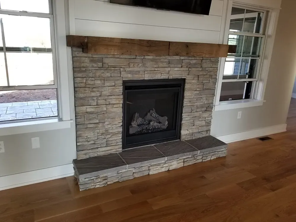 A stone fireplace with a wooden mantel, a tiled hearth, and shiplap paneling above, positioned between two windows.