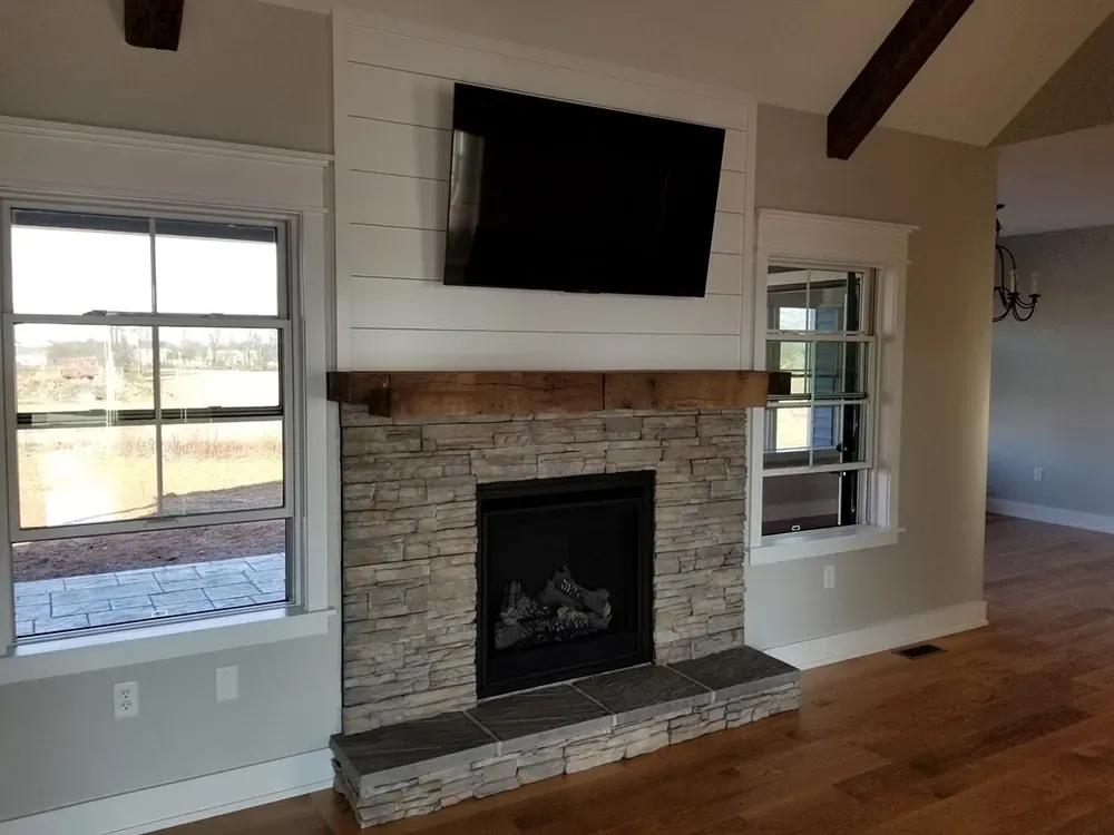 A stone fireplace with a wooden mantel and white shiplap wall, featuring a mounted TV, flanked by two large windows.
