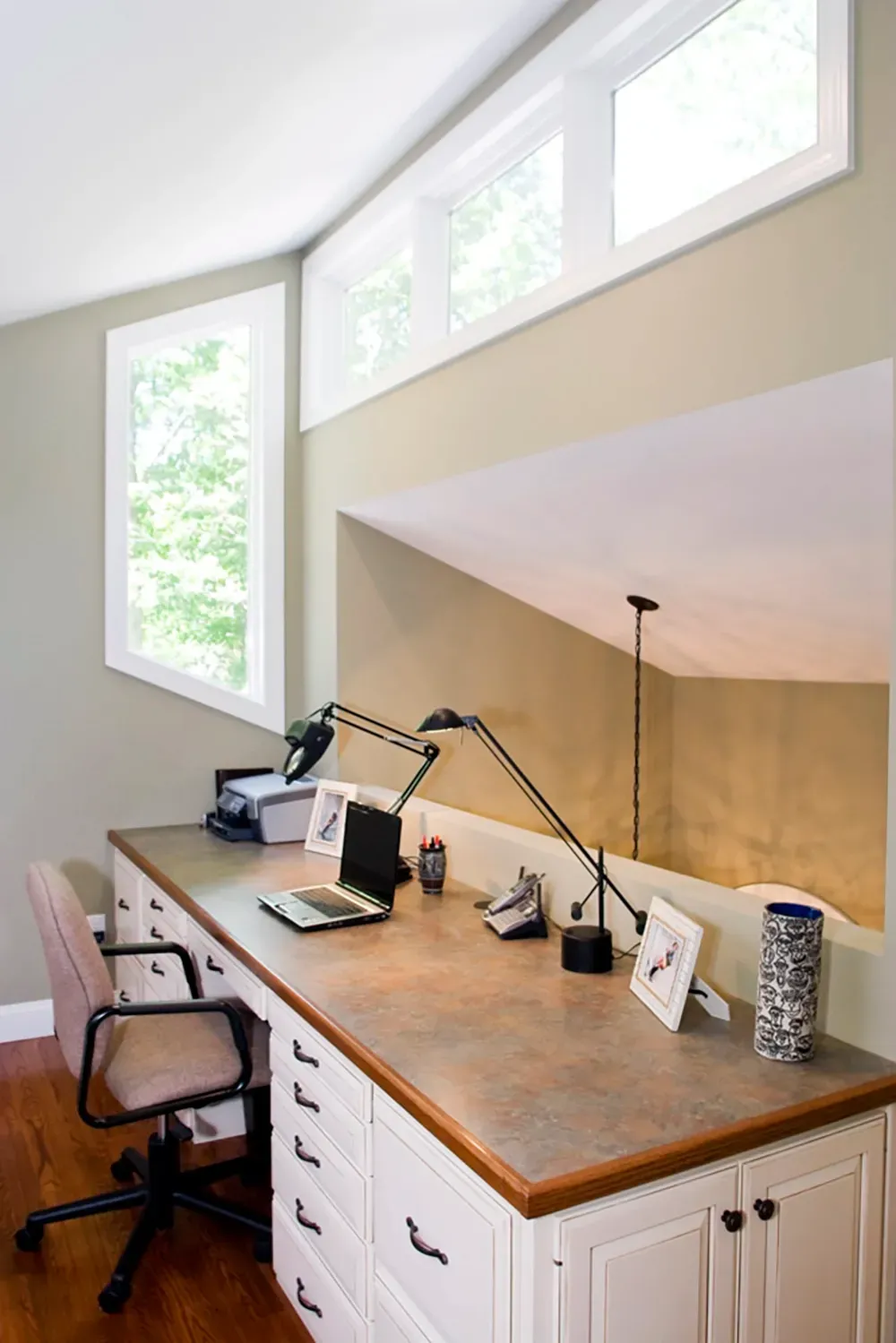A home office with a long desk, white cabinets, and an office chair, set under slanted ceilings and large windows.