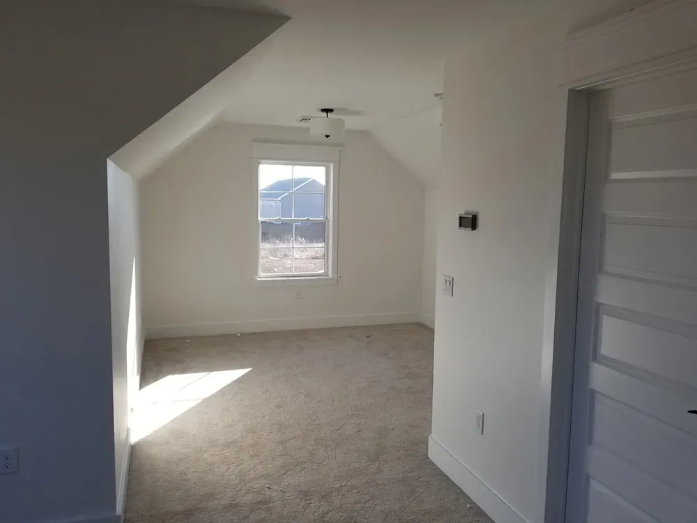 A bright, empty attic room with white walls, beige carpeting, a single window, and a white interior door.