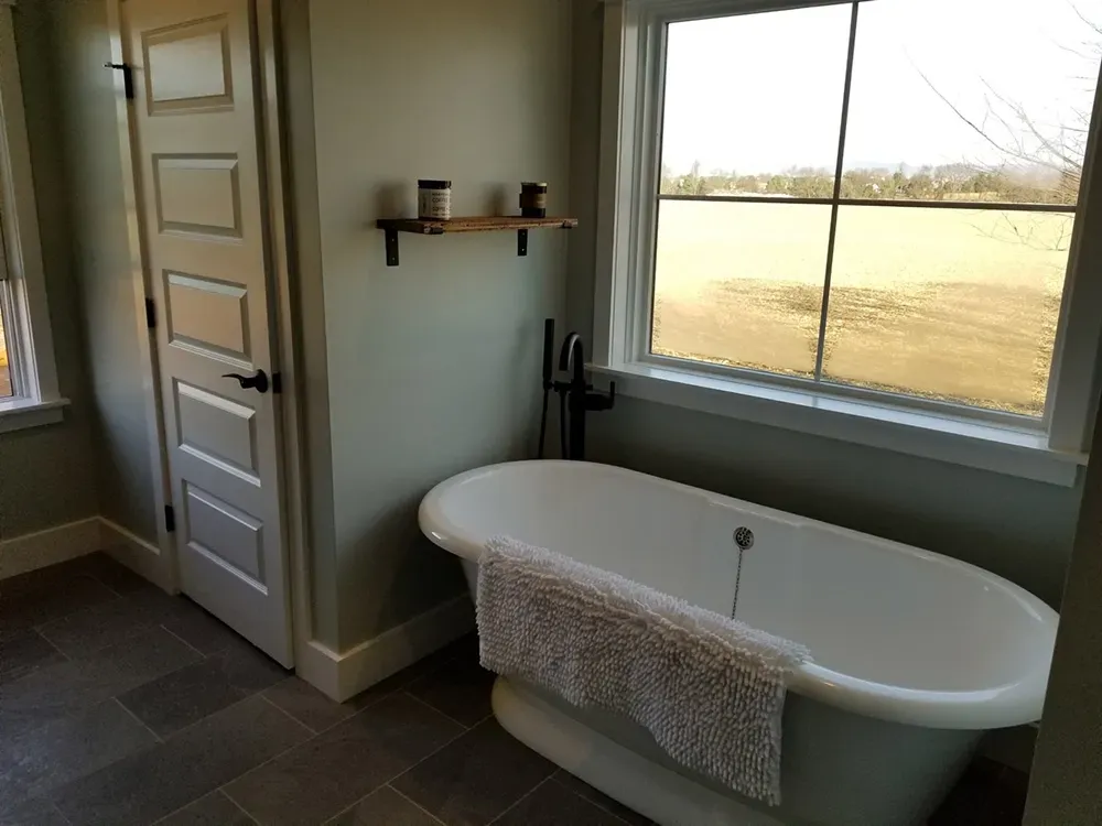 A white freestanding bathtub sits by a large window in a bathroom with pale green walls and dark gray floor tiles.