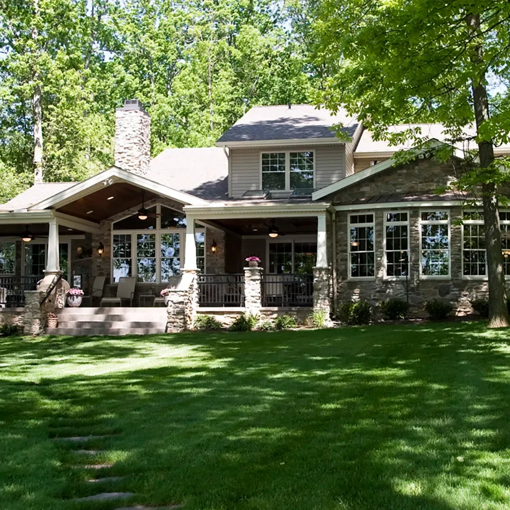 A spacious two-story house with stone accents and a covered back patio, surrounded by lush trees and a green lawn.