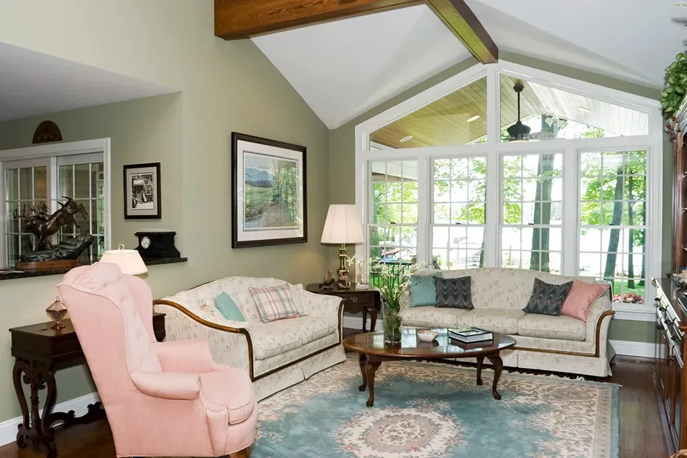 Living room with a pink armchair, two cream sofas, wooden coffee table, blue patterned rug, and large windows with trees.
