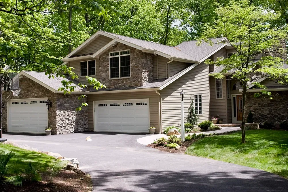 A two-story tan house with a stone facade, two garage doors, and a paved driveway surrounded by lush green trees.