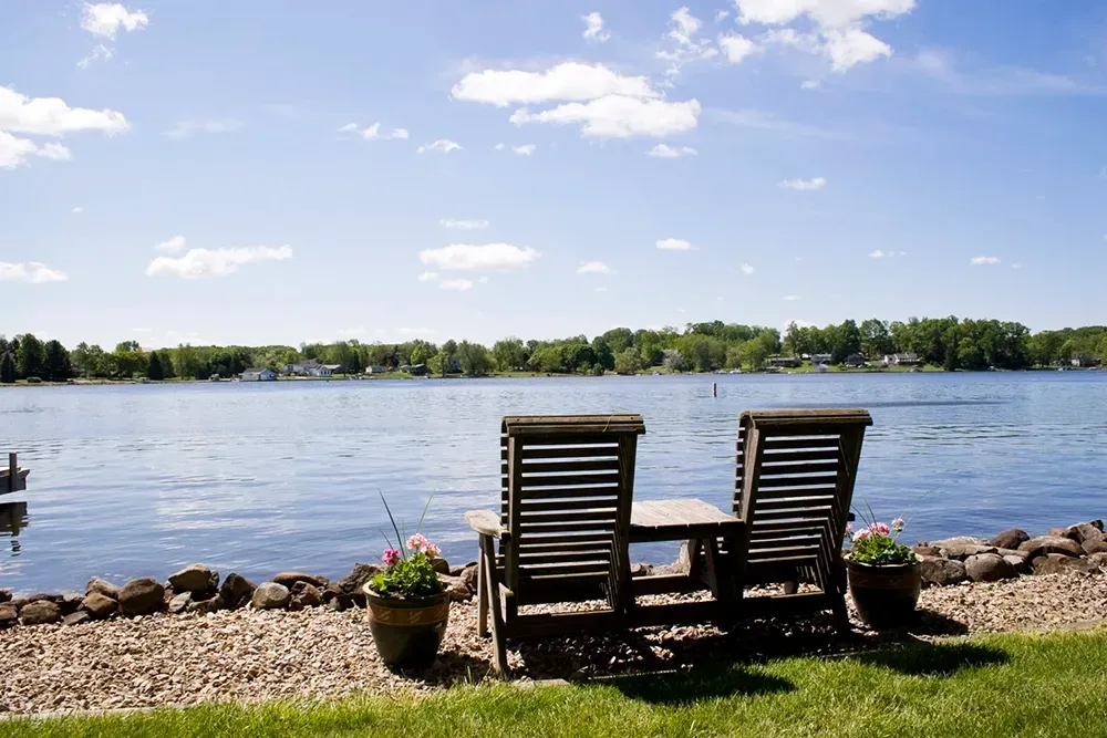 Two wooden chairs and a table sit on a pebble shore overlooking a sunny lake with trees on the opposite bank.