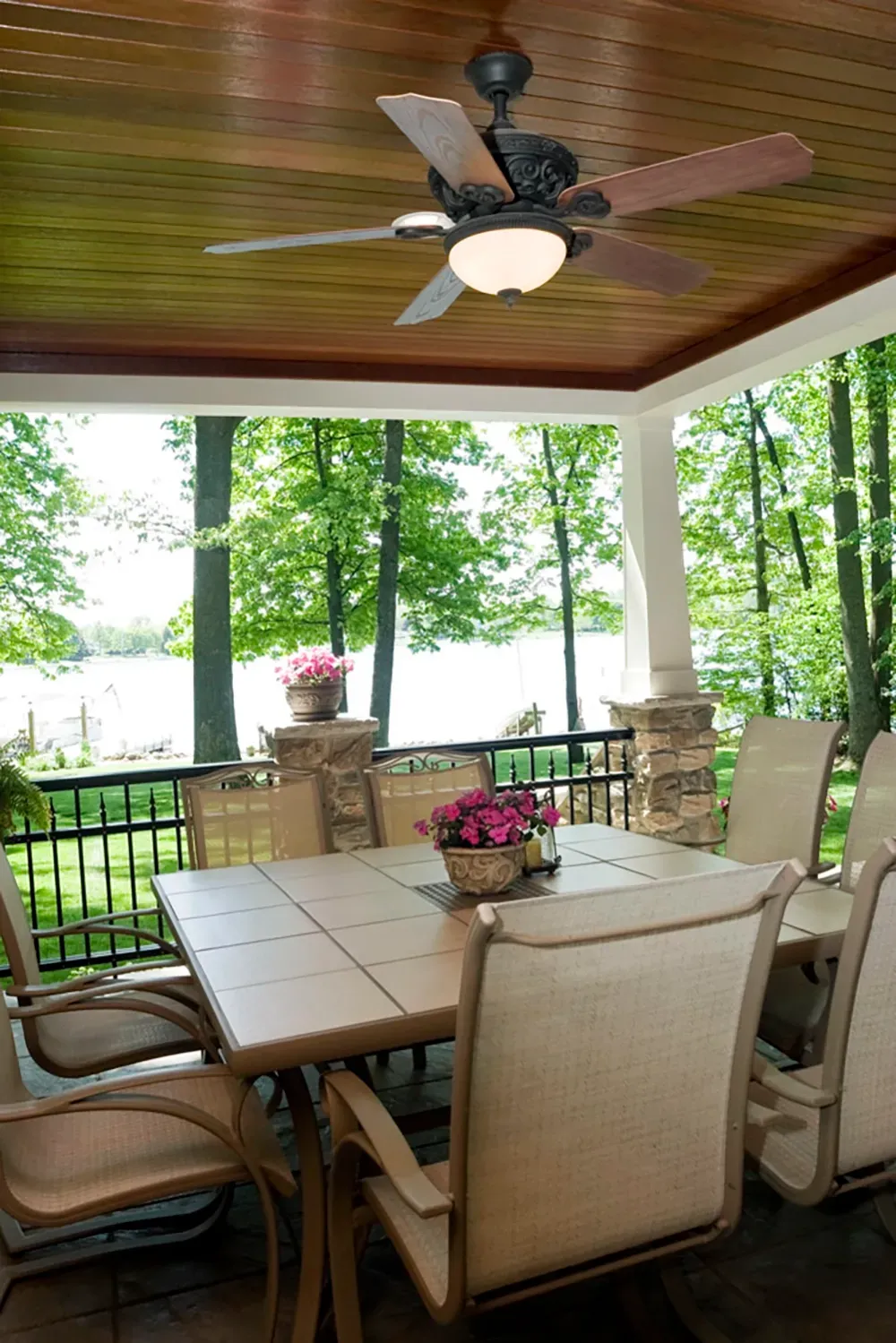 A covered outdoor dining patio with a table and chairs, featuring a wooden ceiling, ceiling fan, and views of a lake.