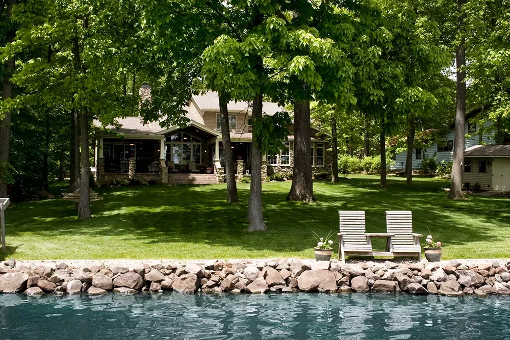 A house with a porch sits behind a grassy lawn and a stone-lined waterfront with two wooden chairs.