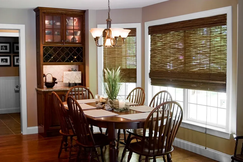 A dining room with a wooden table, chairs, a glass-front cabinet, a chandelier, and windows with bamboo shades.