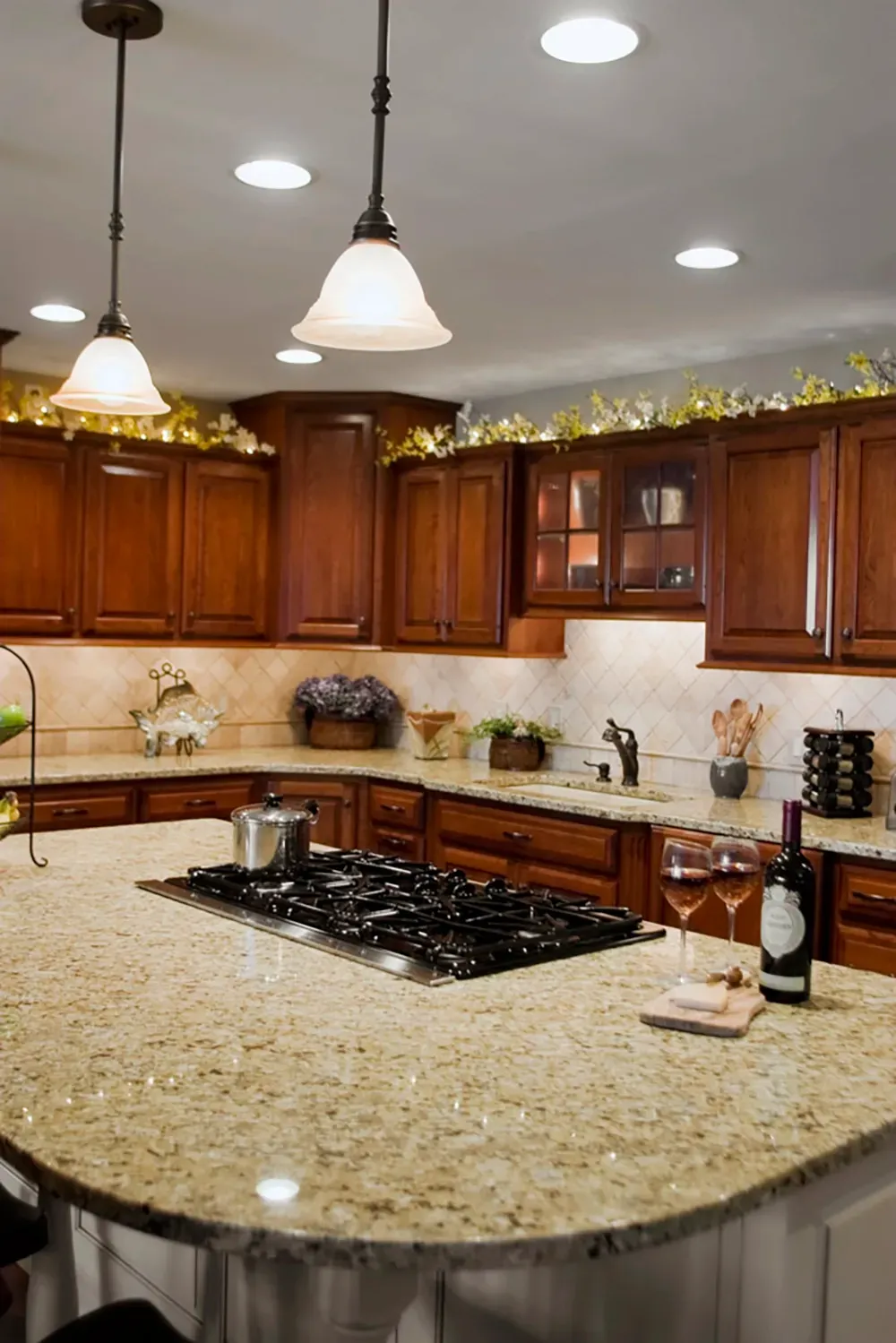 A kitchen island with a black stovetop, granite counters, wood cabinets, and two wine glasses under pendant lighting.