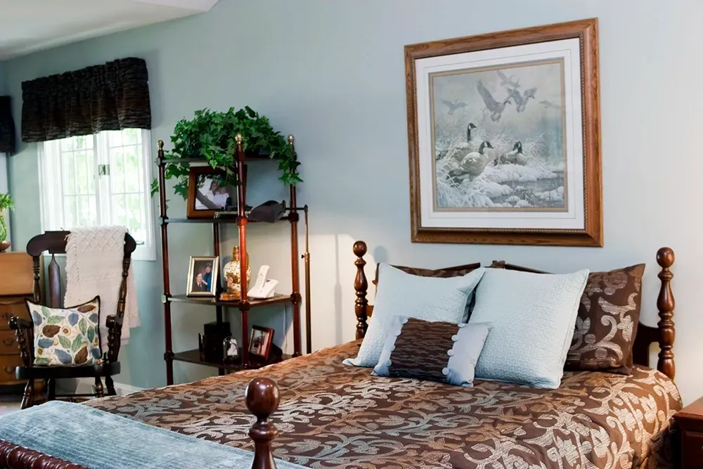A bedroom features a bed with a brown patterned quilt, light blue pillows, and framed wildlife art on a pale blue wall.