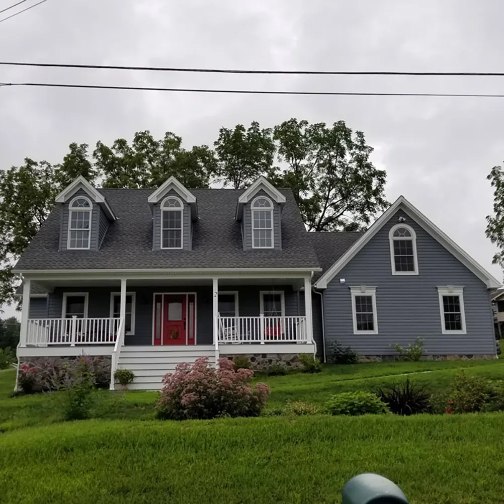 A two-story blue house with a red front door, a white porch, and three dormer windows on a grassy lawn under a cloudy sky.