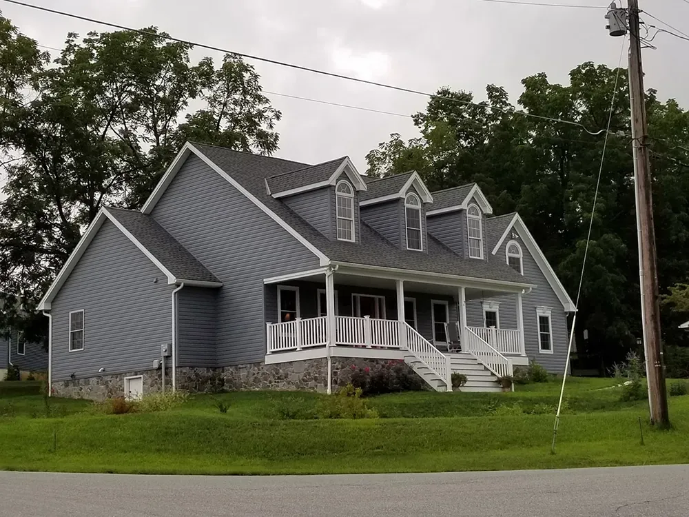 A blue two-story house with a white front porch, stone foundation, and three roof dormers in a grassy, rural setting.