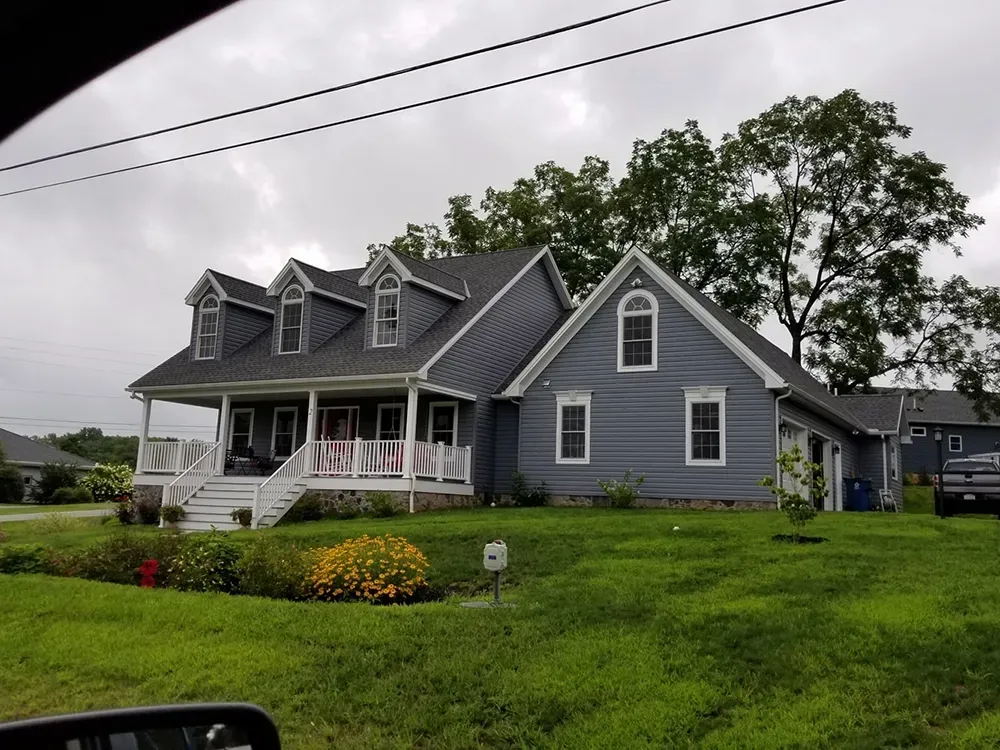A two-story blue house with a white front porch and three dormer windows, surrounded by a green lawn and trees.