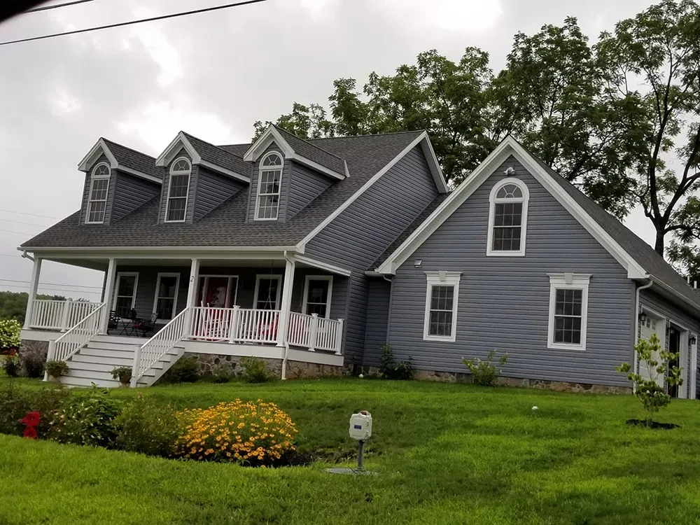 A blue, two-story house with a front porch, three dormer windows, and a lush green lawn under a cloudy sky.
