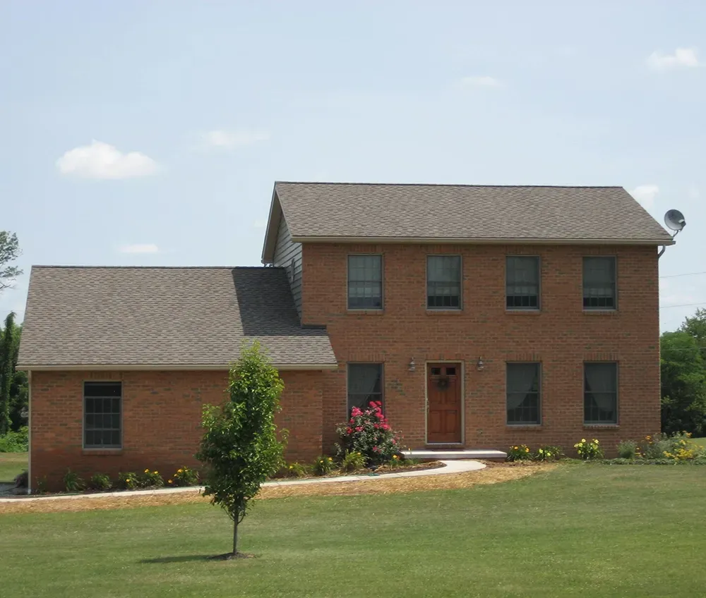 A two-story red brick house with a brown shingled roof, a front door, and green lawn, set against a blue sky.