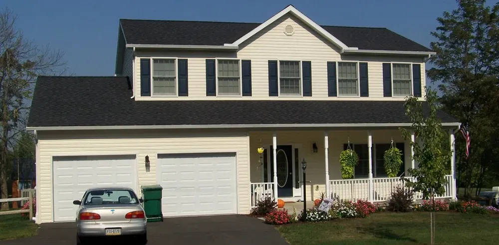 A two-story, cream-colored suburban house with a dark shingled roof, two-car garage, and a front porch with a parked car.