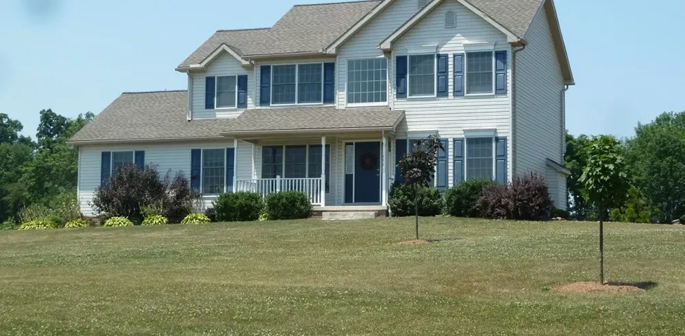 A two-story, white suburban house with blue shutters, a brown shingled roof, and a large front lawn under a clear blue sky.
