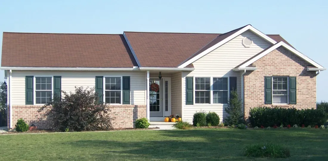 A one-story ranch-style house with beige siding, brick accents, brown roof shingles, and green shutters in a grassy yard.