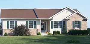 A one-story suburban home with beige siding, brick accents, a brown roof, and dark green shutters on a grassy lawn.