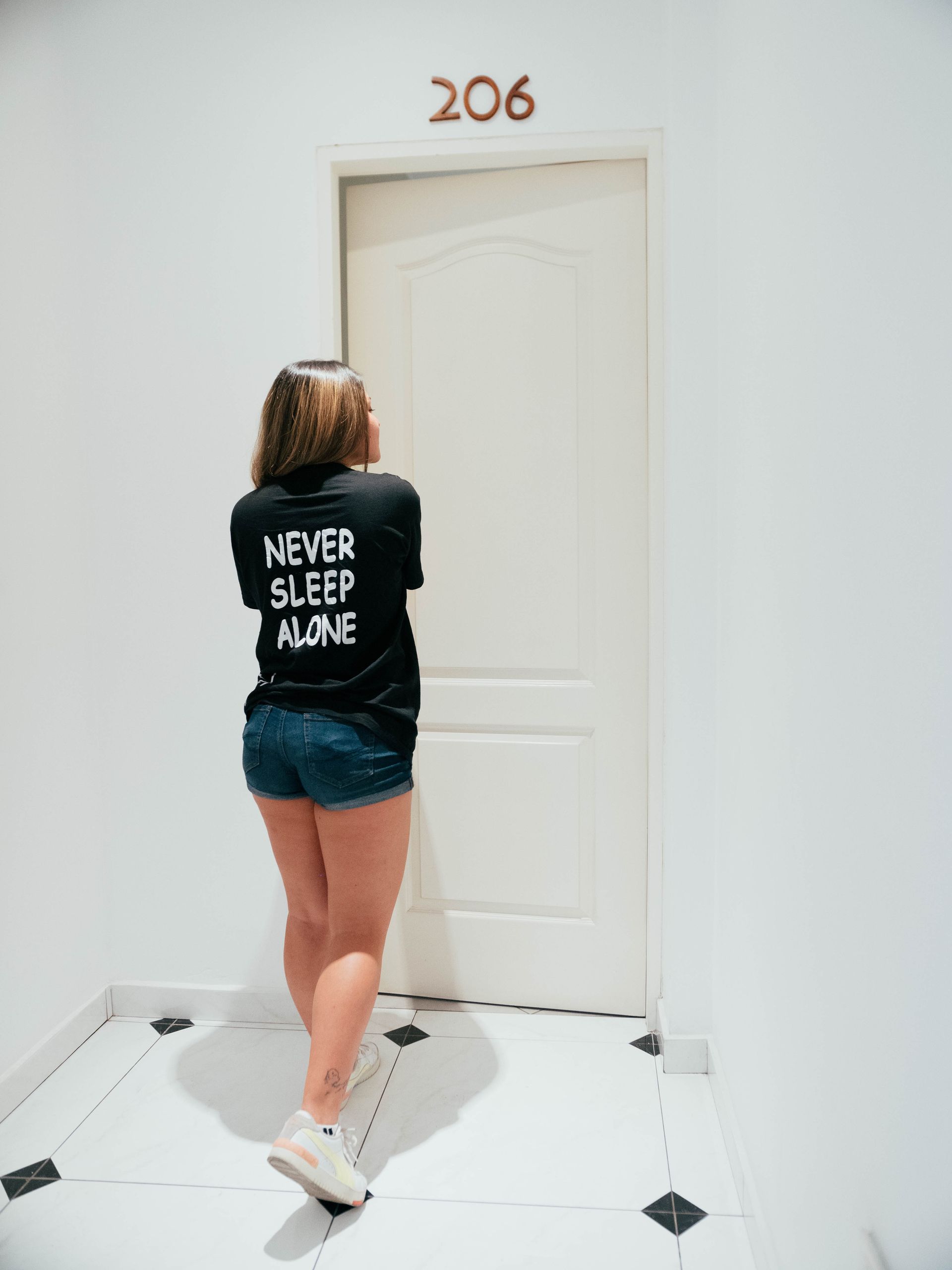 A woman wearing a black shirt that says `` never sleep alone '' is standing in front of a door.