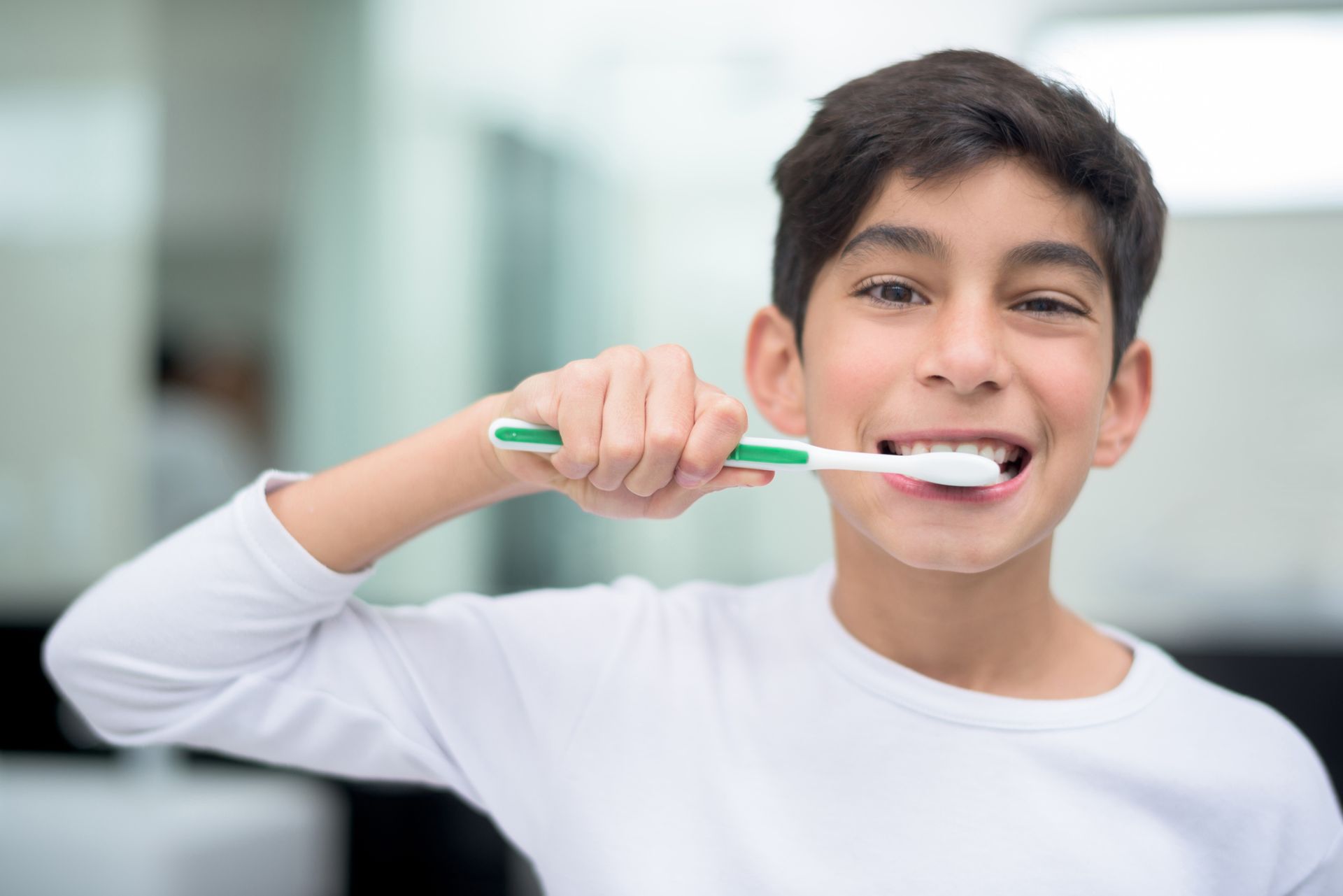 Boy brushes teeth with a green and white toothbrush in a bathroom, smiling.