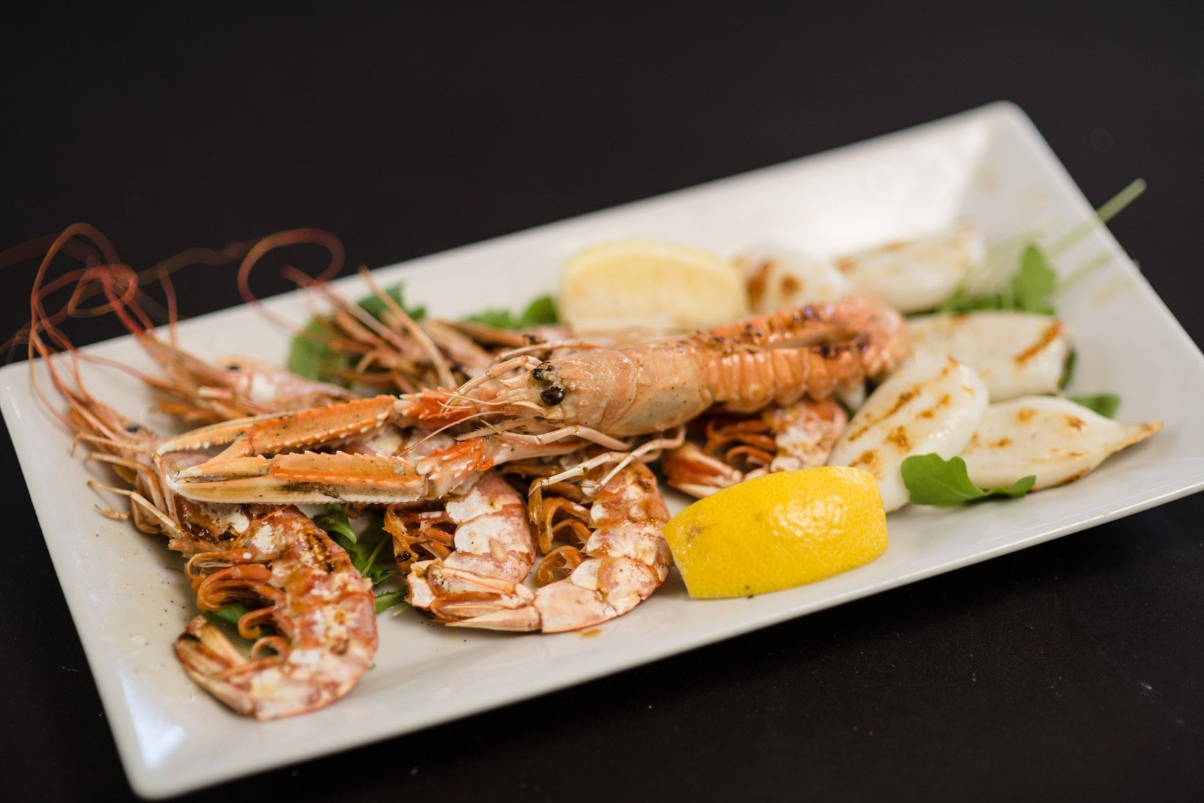 Piatto di pesce alla griglia: gamberi, calamari, spicchi di limone e verdure su un piatto rettangolare bianco.