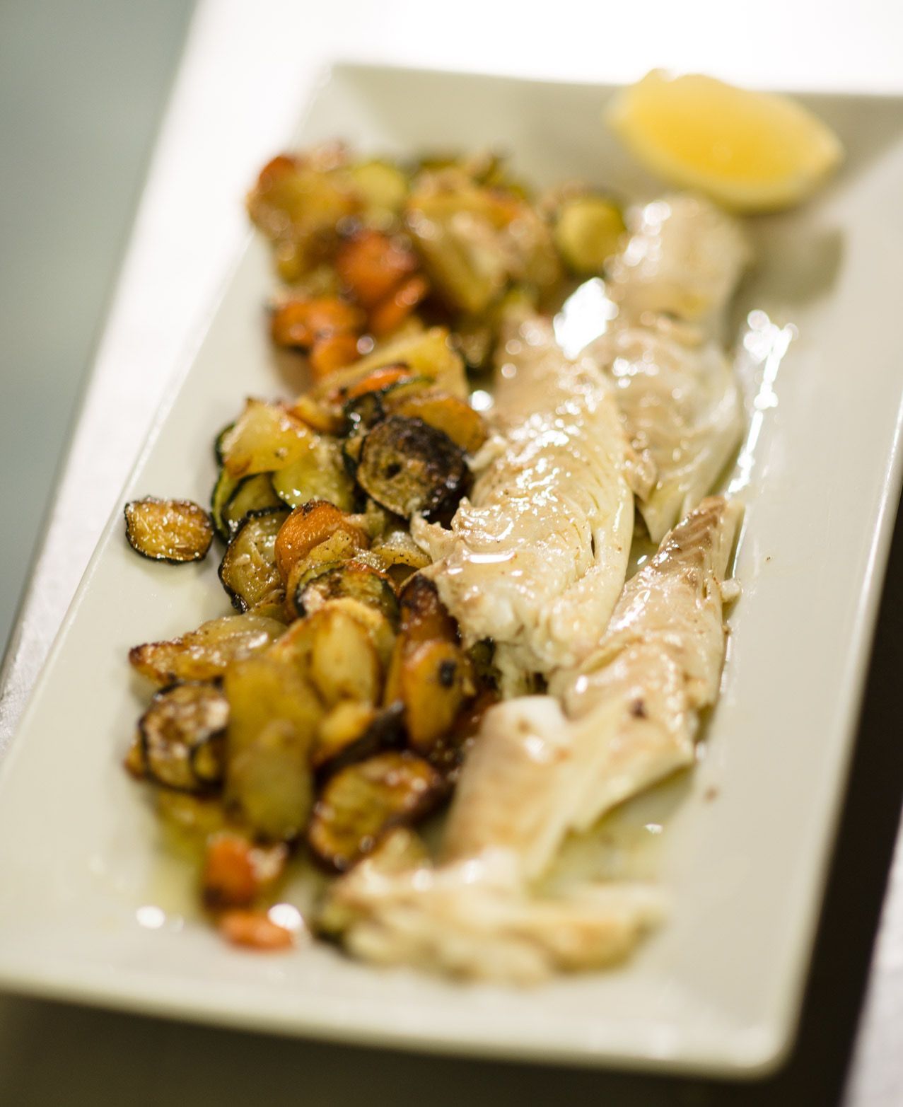 Filetti di pesce con verdure arrostite e uno spicchio di limone su un piatto bianco rettangolare.