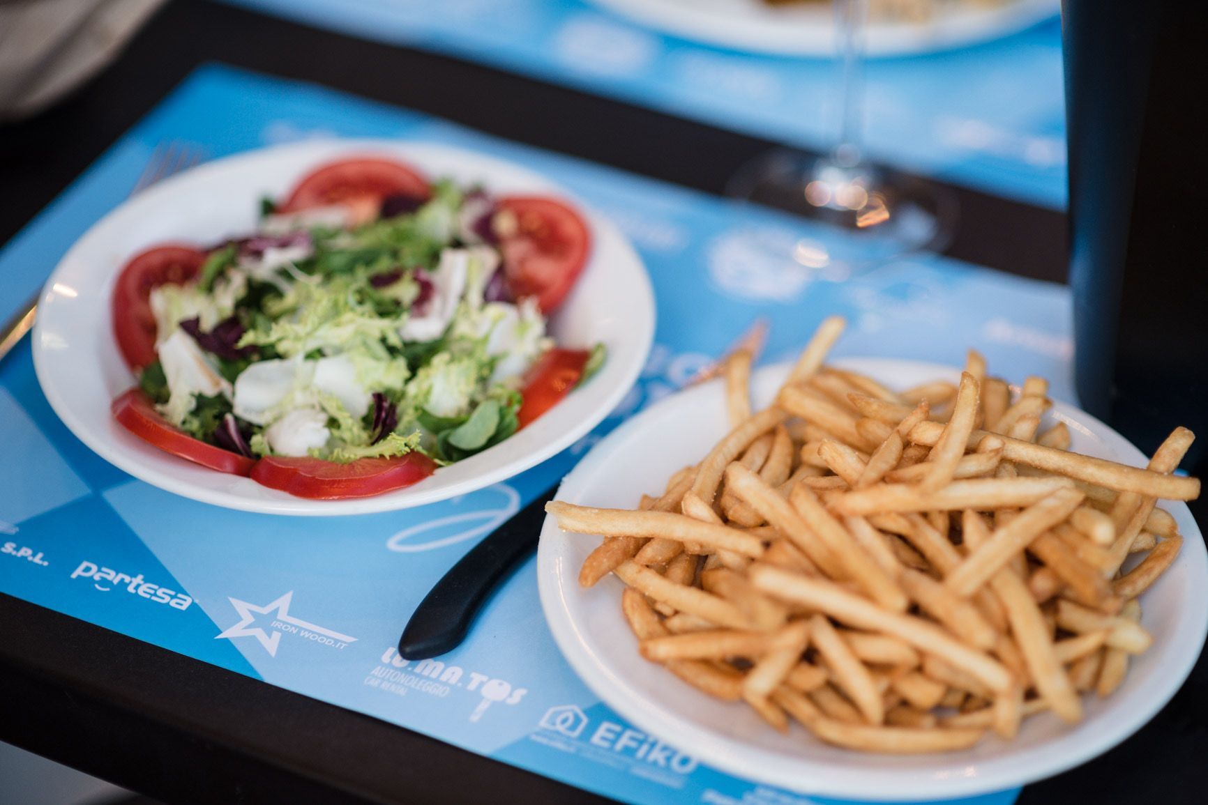 Insalata e patatine fritte su piatti bianchi sopra una tovaglietta blu, in un ristorante all'aperto.