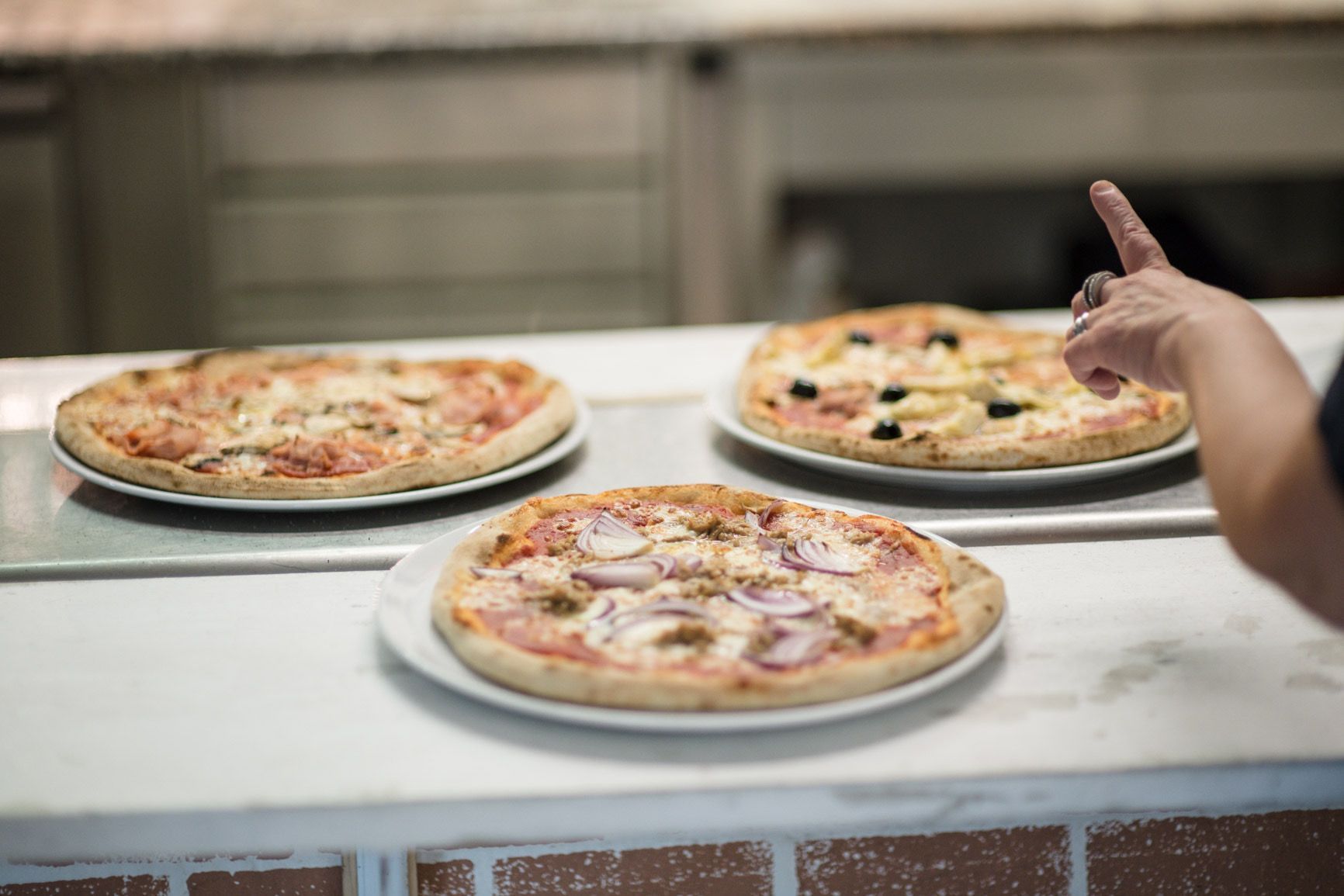 Tre pizze su piatti bianchi al bancone, una delle quali è indicata dalla mano di una persona.