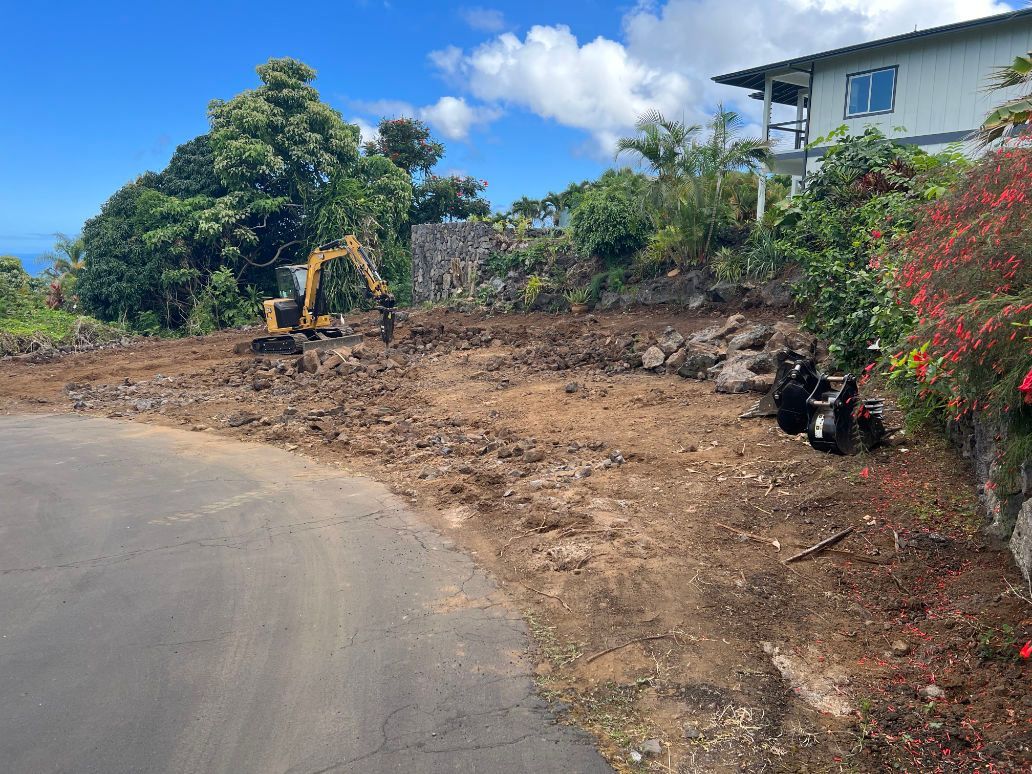 A house is being built on a hill next to a dirt road showing a excavator. 