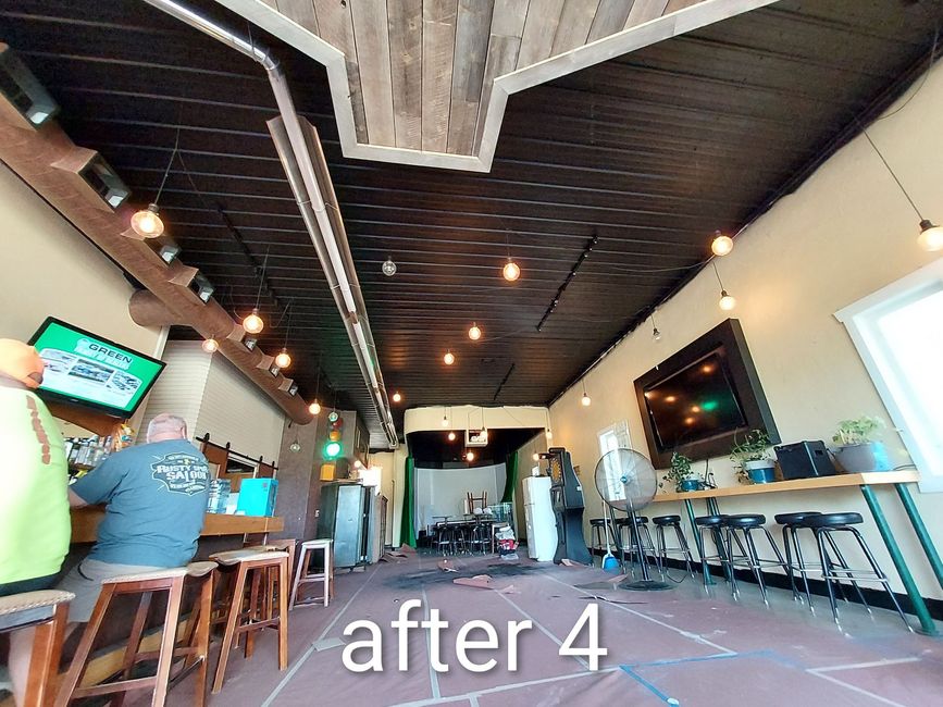 Wide-angle view of a rustic bar interior with high stools, TV screens, and a few patrons sitting at the counter