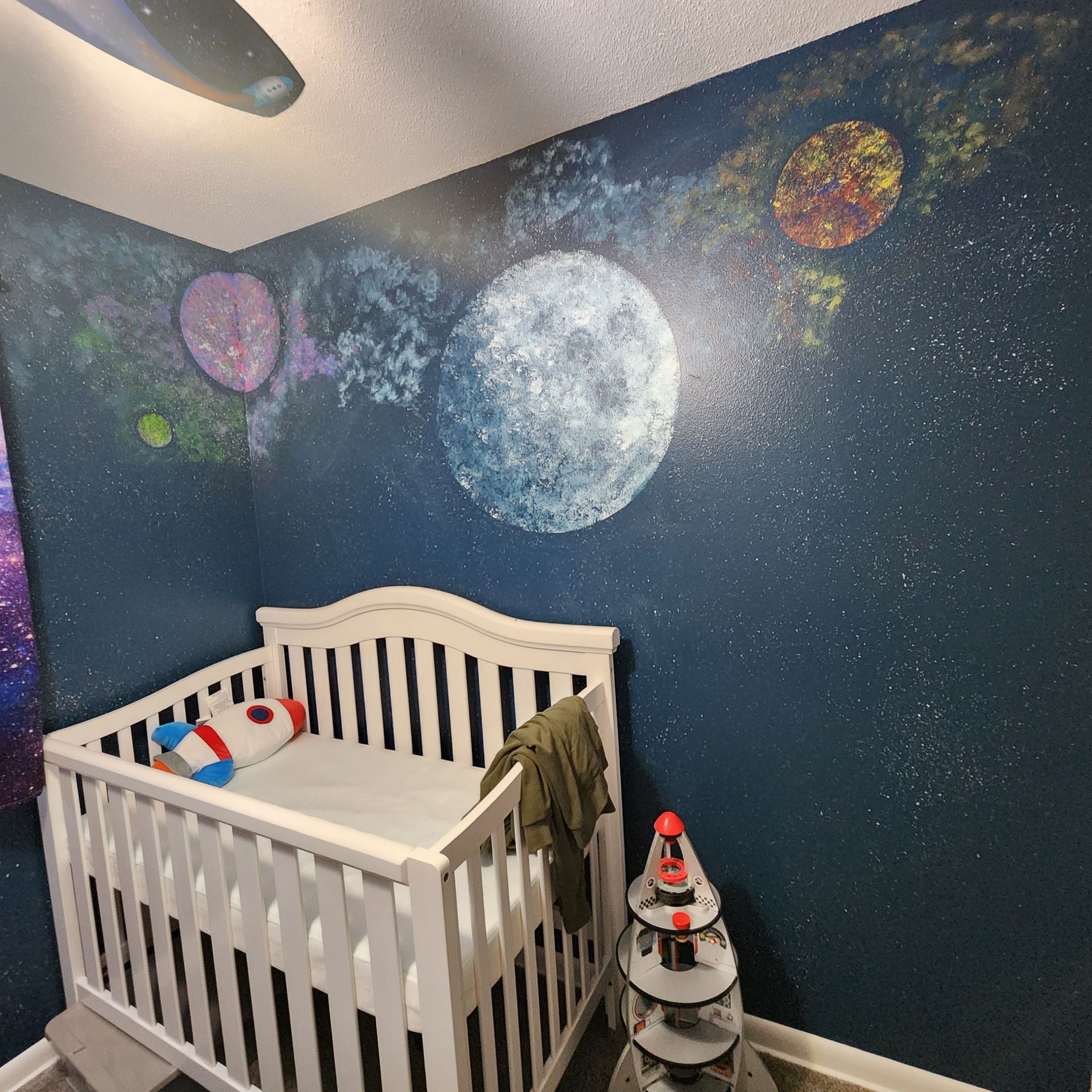White crib in a starry space-themed nursery with painted planets and moon on dark blue walls.