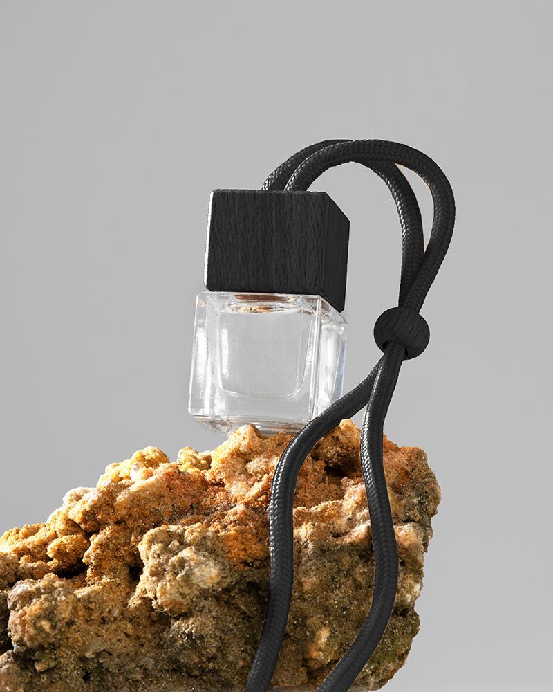 autoparfum helder glas met zwart snoer, hangend boven een ruwe rots op een grijze achtergrond.