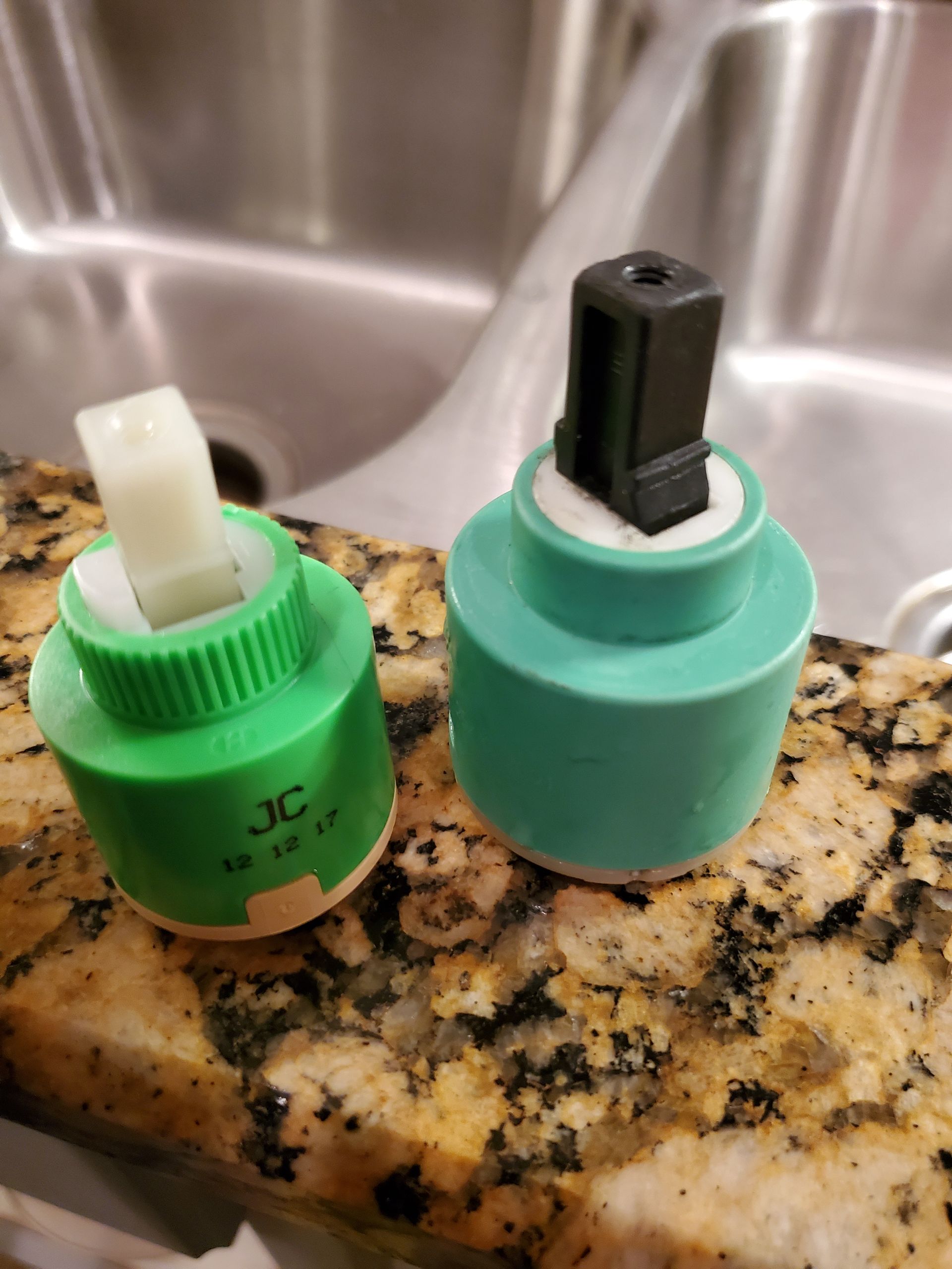Two ceramic faucet cartridges, green and teal, with white and black parts, on a marble countertop.
