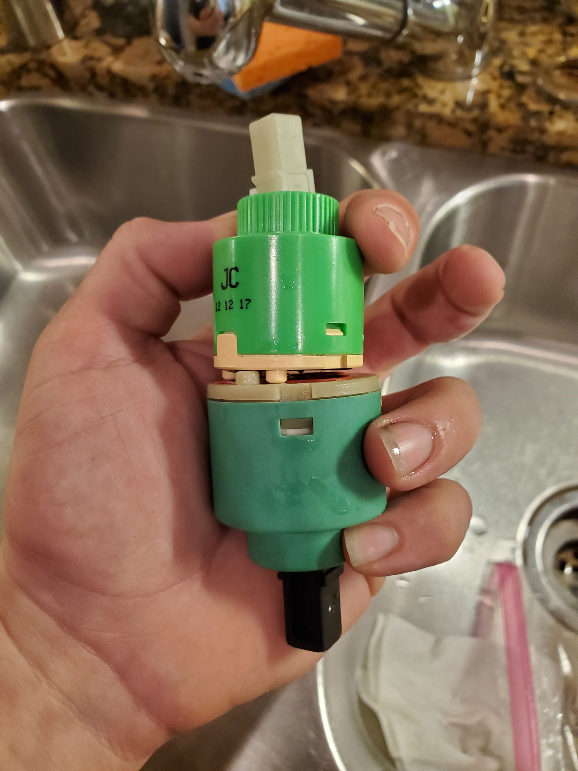 Hand holding a green and beige faucet cartridge over a stainless steel sink.