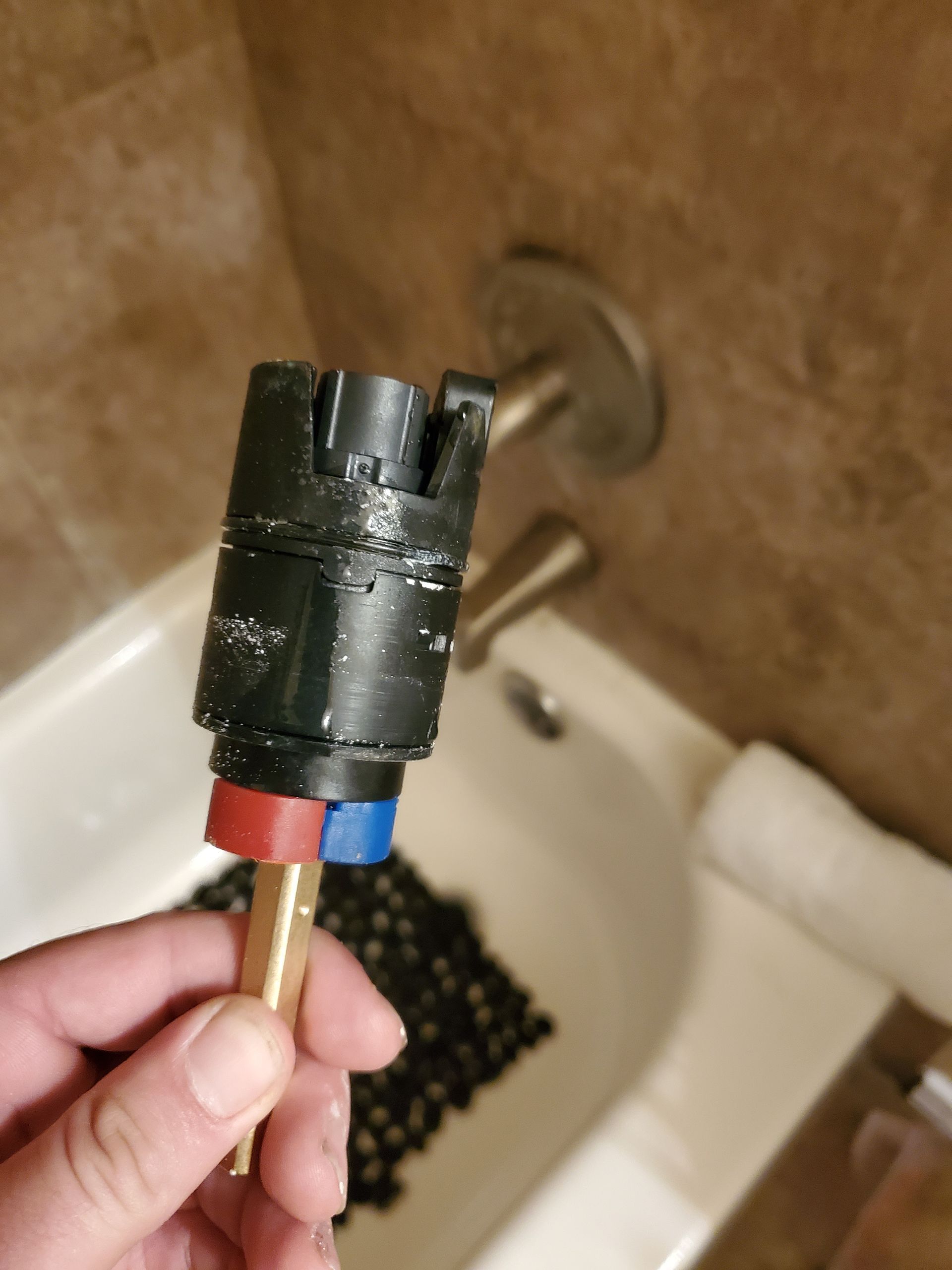A person holding a black valve cartridge with red/blue color-coded ends, in front of a bathtub faucet.