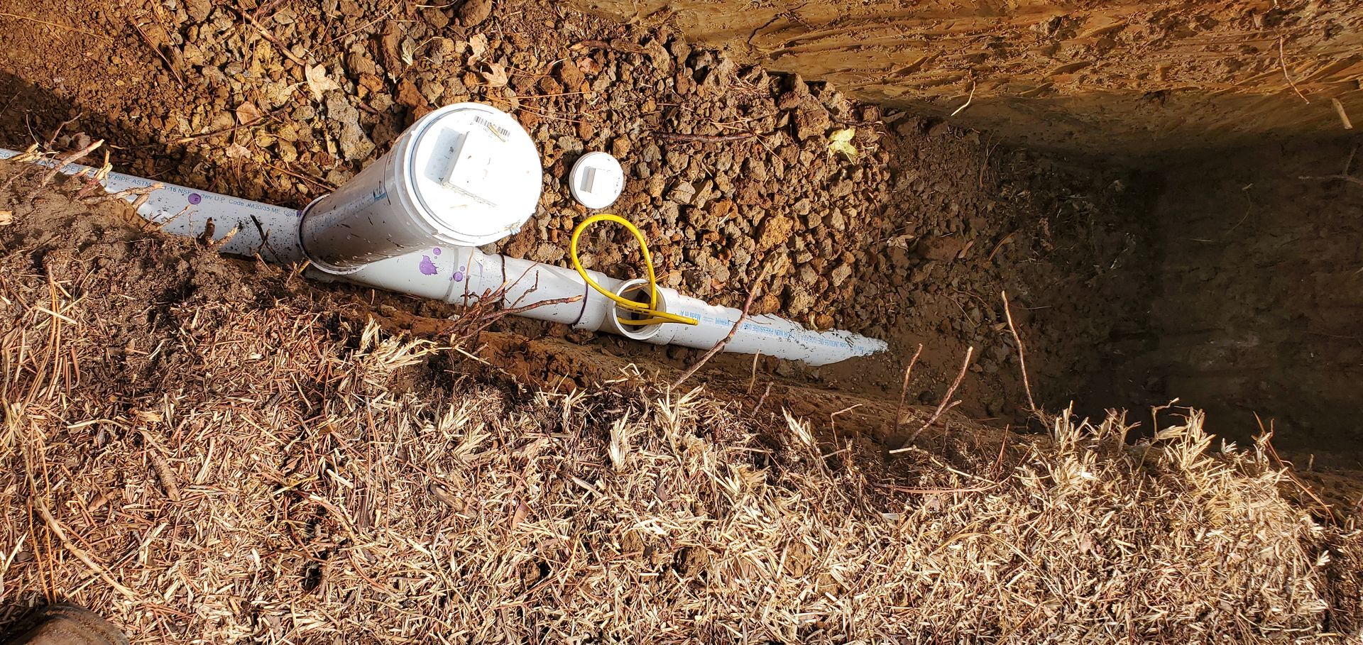 PVC pipes in a trench. One pipe has a cap. Soil and mulch are present.