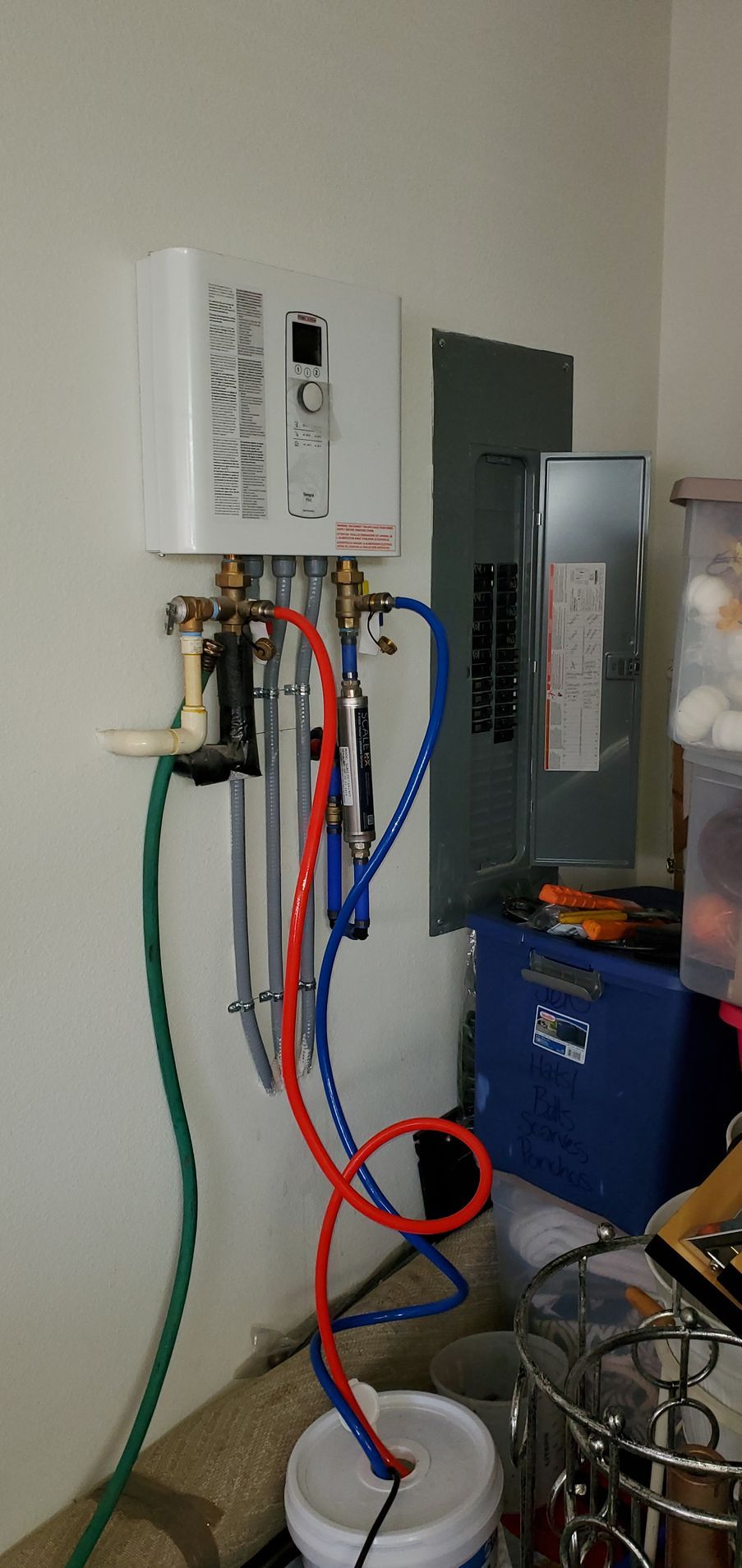 White tankless water heater, connected with red/blue pipes and green hose, next to a breaker box.