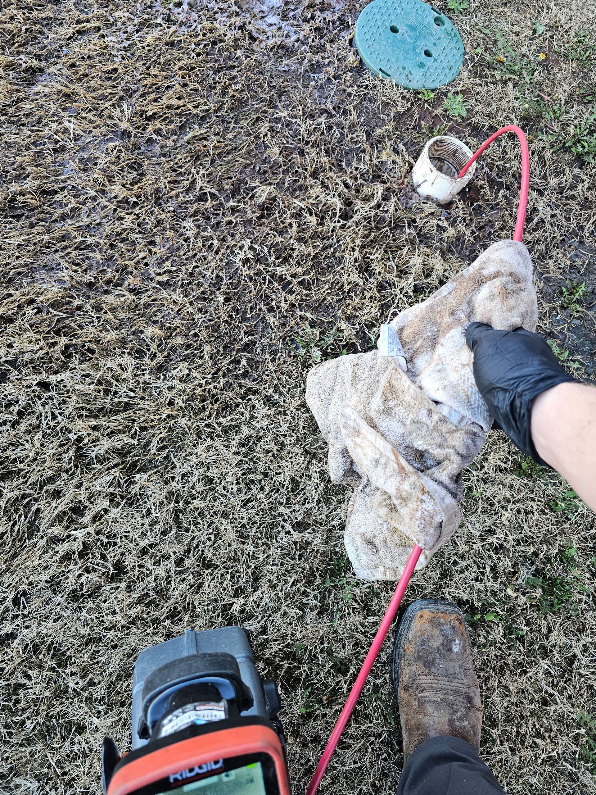 A gloved hand pulls debris from a drain line. A camera, hose, and drain cap sit nearby on mulch.