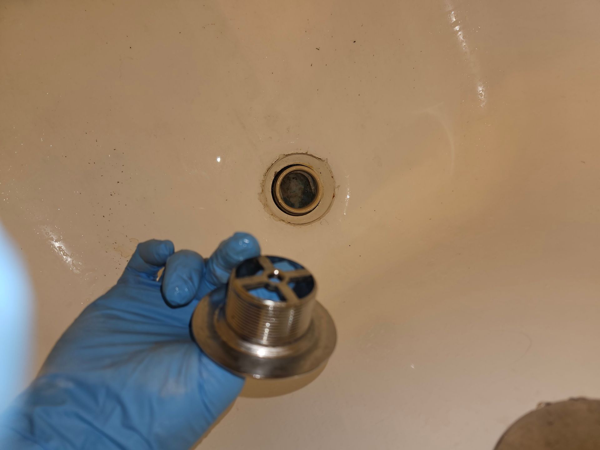 Hand in blue glove holding a bathtub drain, next to a bathtub drain hole.