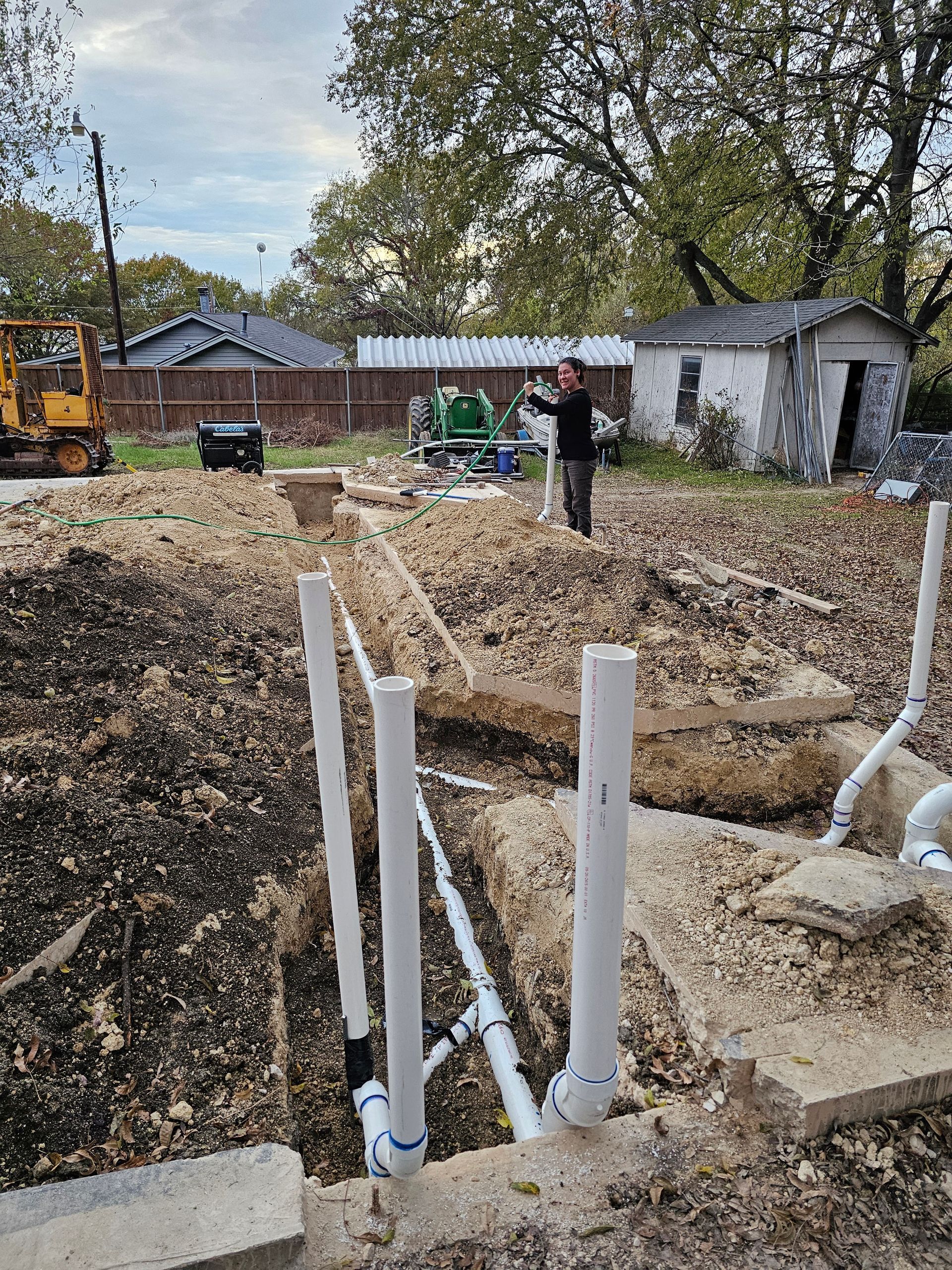 Construction site with exposed pipes. Person standing amidst dirt, trees, and equipment.