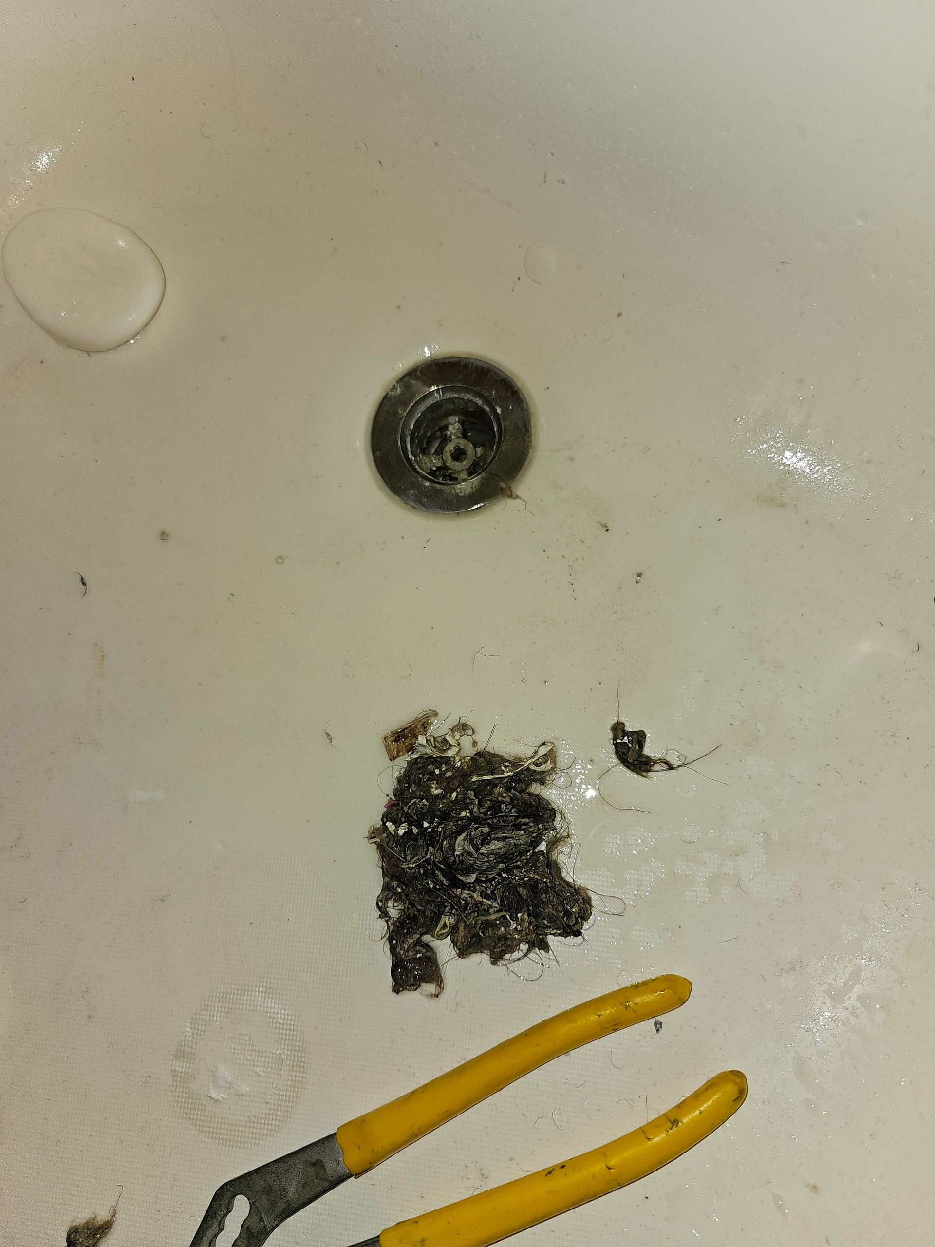 A clogged bathtub drain with a large clump of hair and debris, yellow-handled pliers nearby.