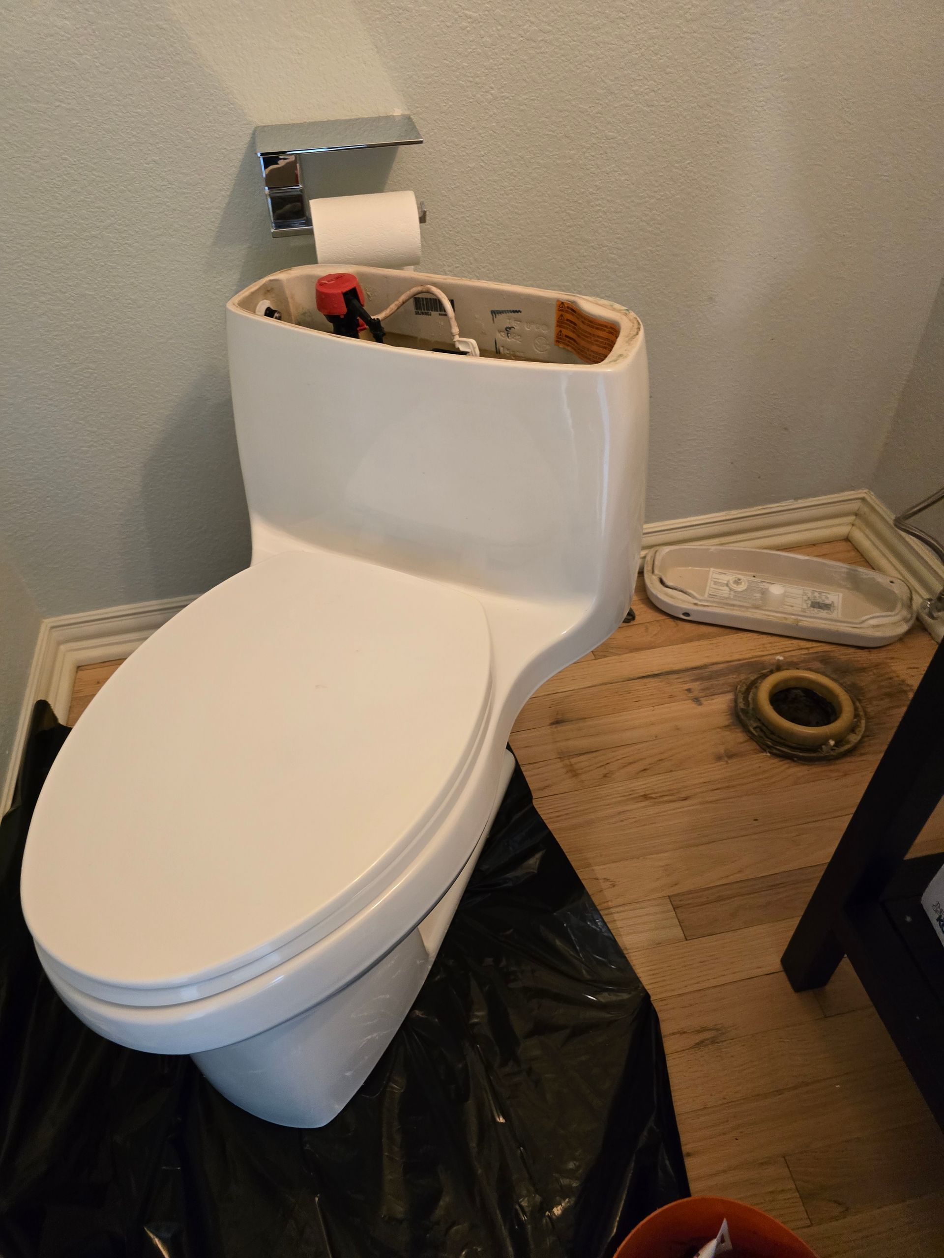 White toilet with tank removed, sitting on plastic, surrounded by wood pieces and plumbing parts.