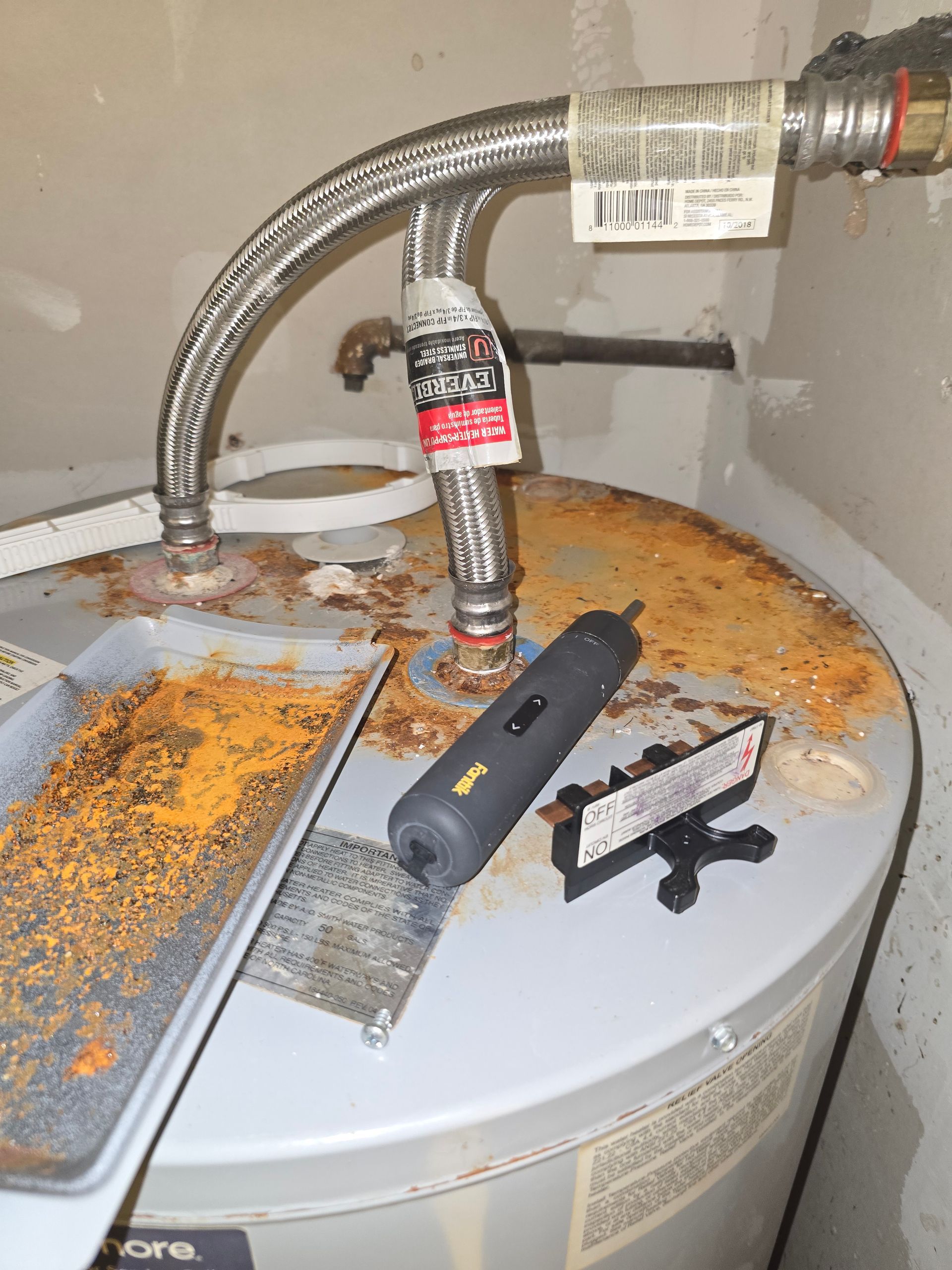 A rusty water heater with a tray of rust debris, tools on top, and metal piping.