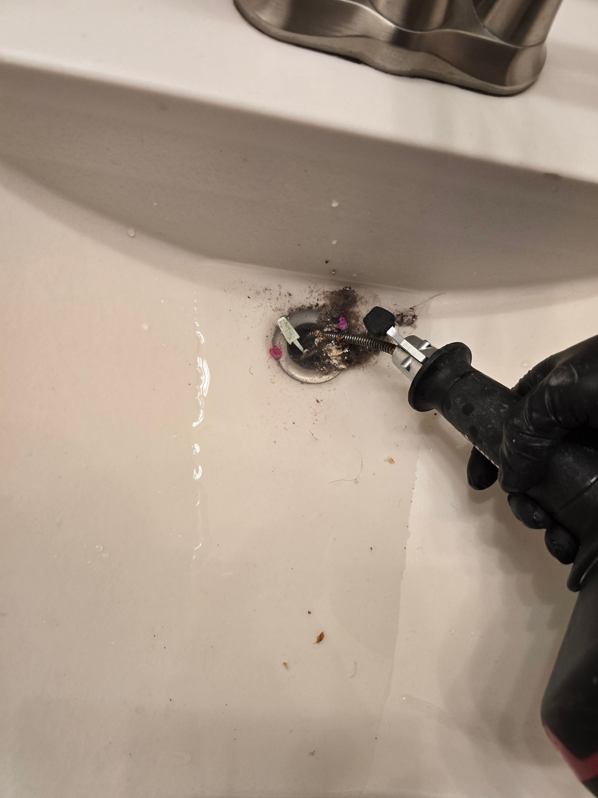 A gloved hand uses a drain cleaner tool in a white sink, removing hair and debris from the drain.