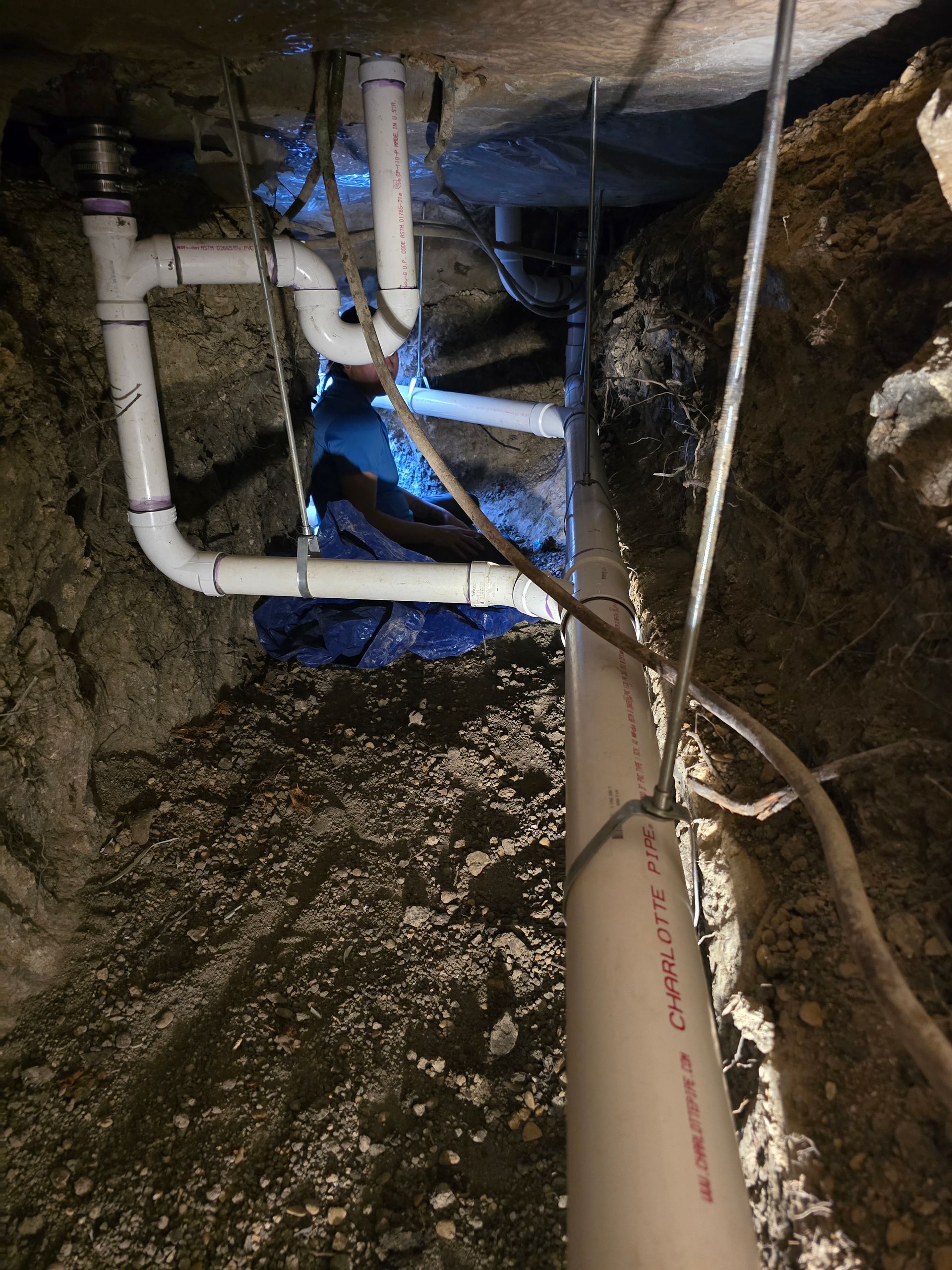 White PVC pipes installed in a narrow trench. Dirt and exposed foundation surround the pipes.