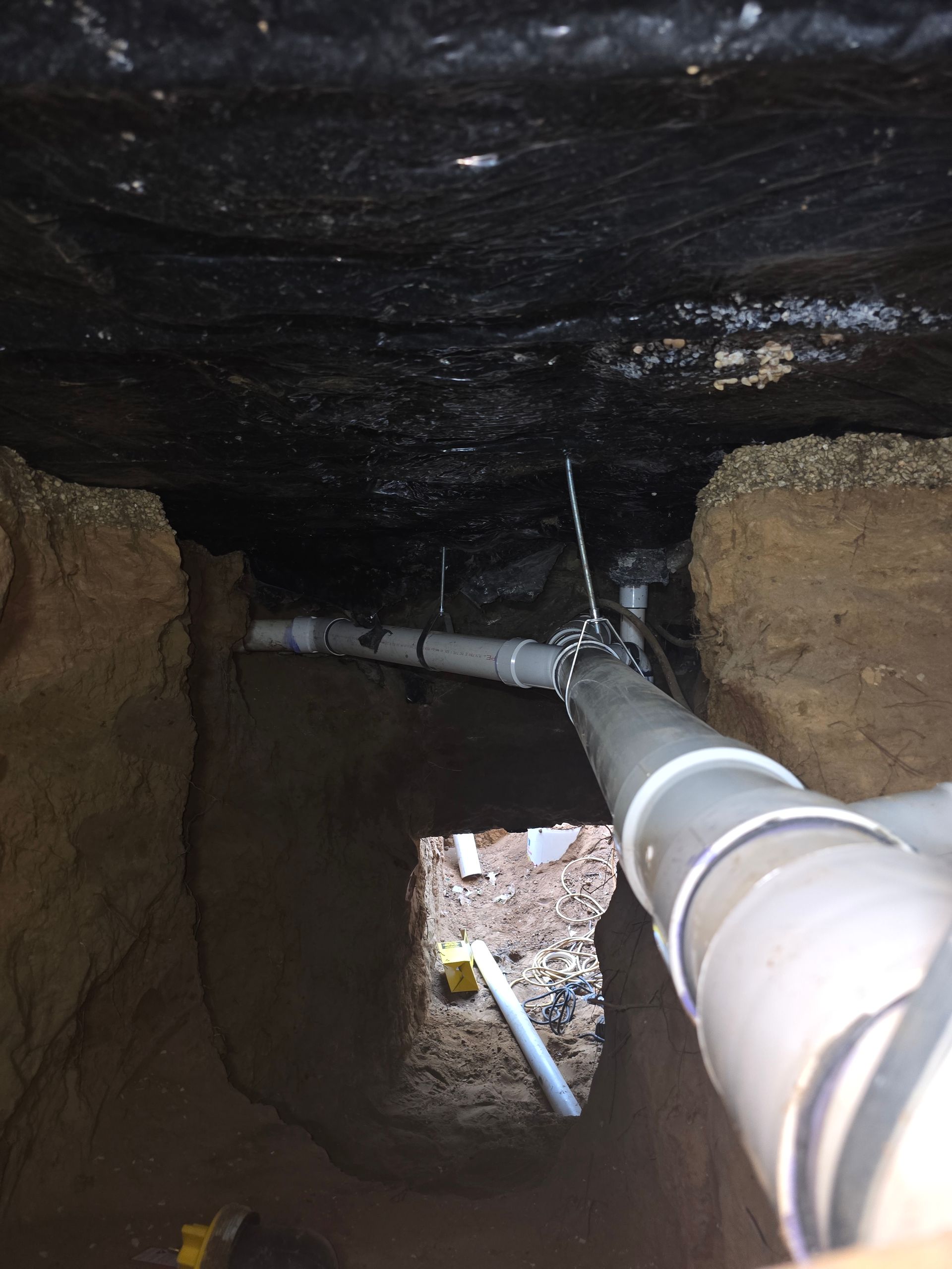Pipes emerging from a dark, enclosed space. Grey PVC pipes connect with white and metal fixtures in a dirt trench.