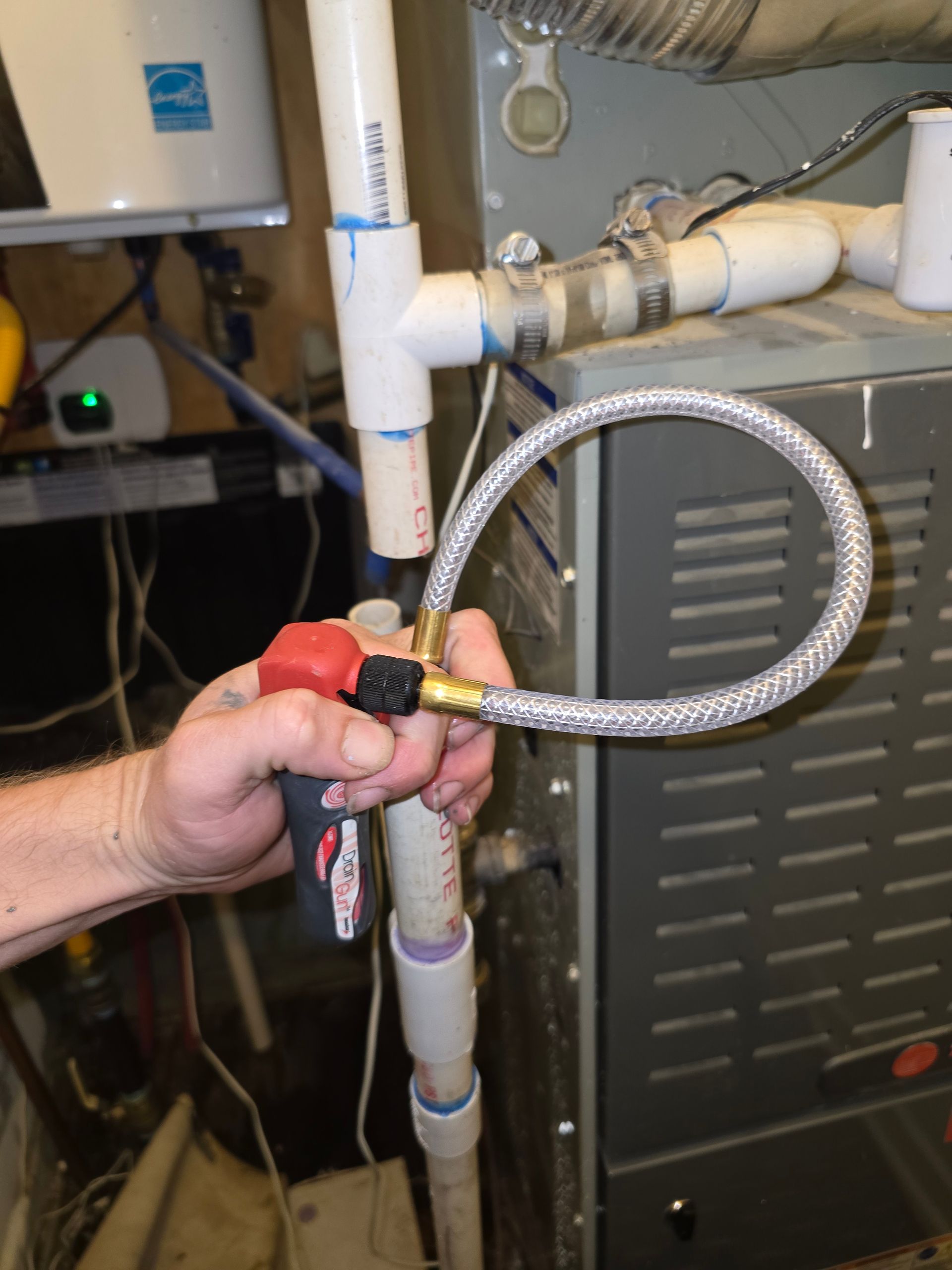 Hand holding a red valve connected by a flexible hose to a PVC pipe near a furnace.