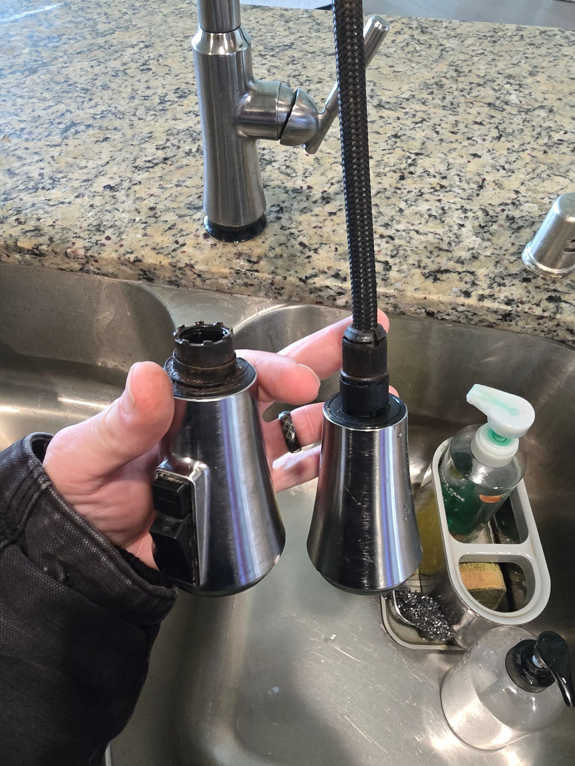 Hand holding two detached faucet spray heads near a sink with granite countertop.