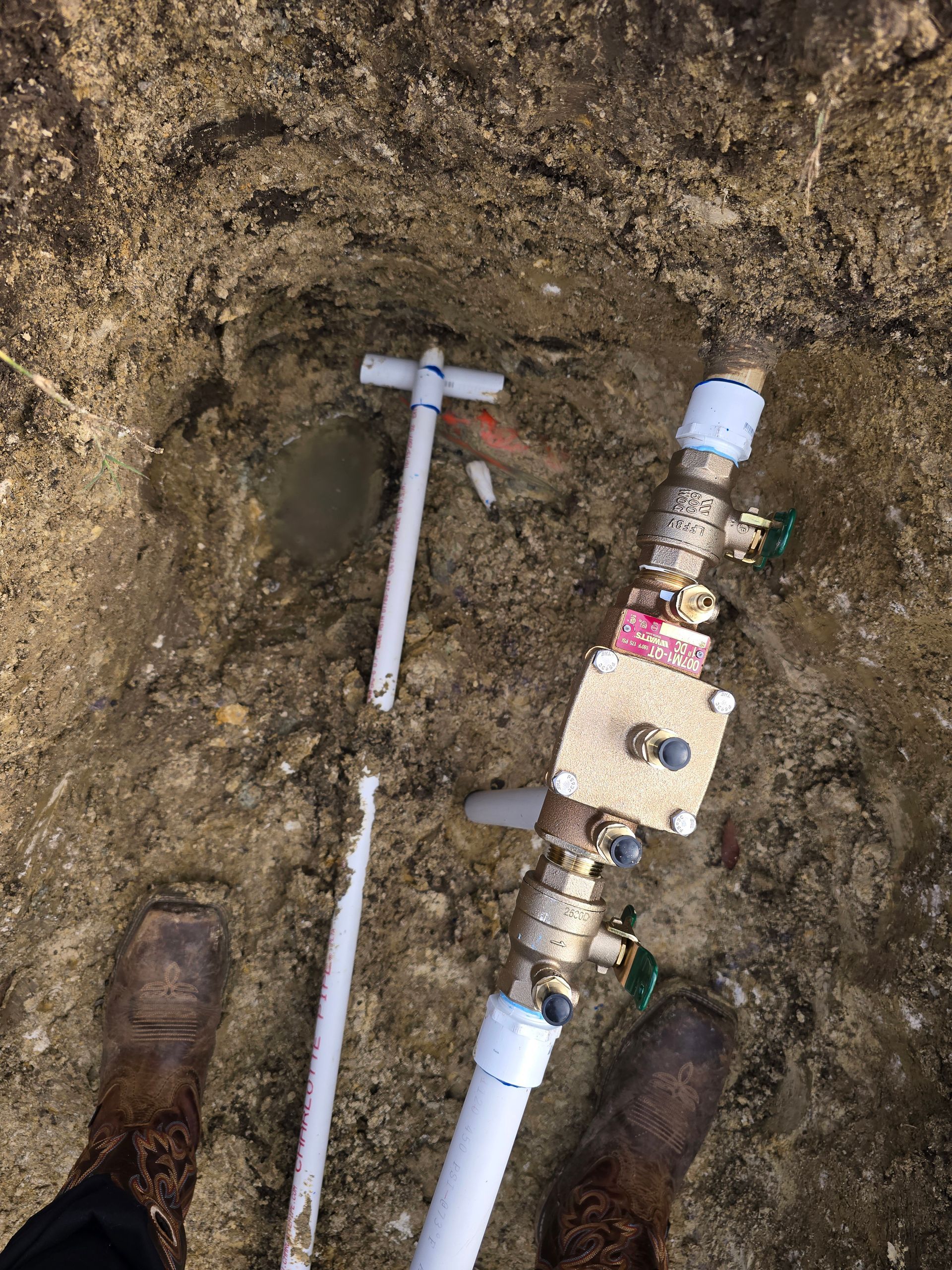 Plumbing components in dirt pit with boots, PVC pipe, and brass valves.